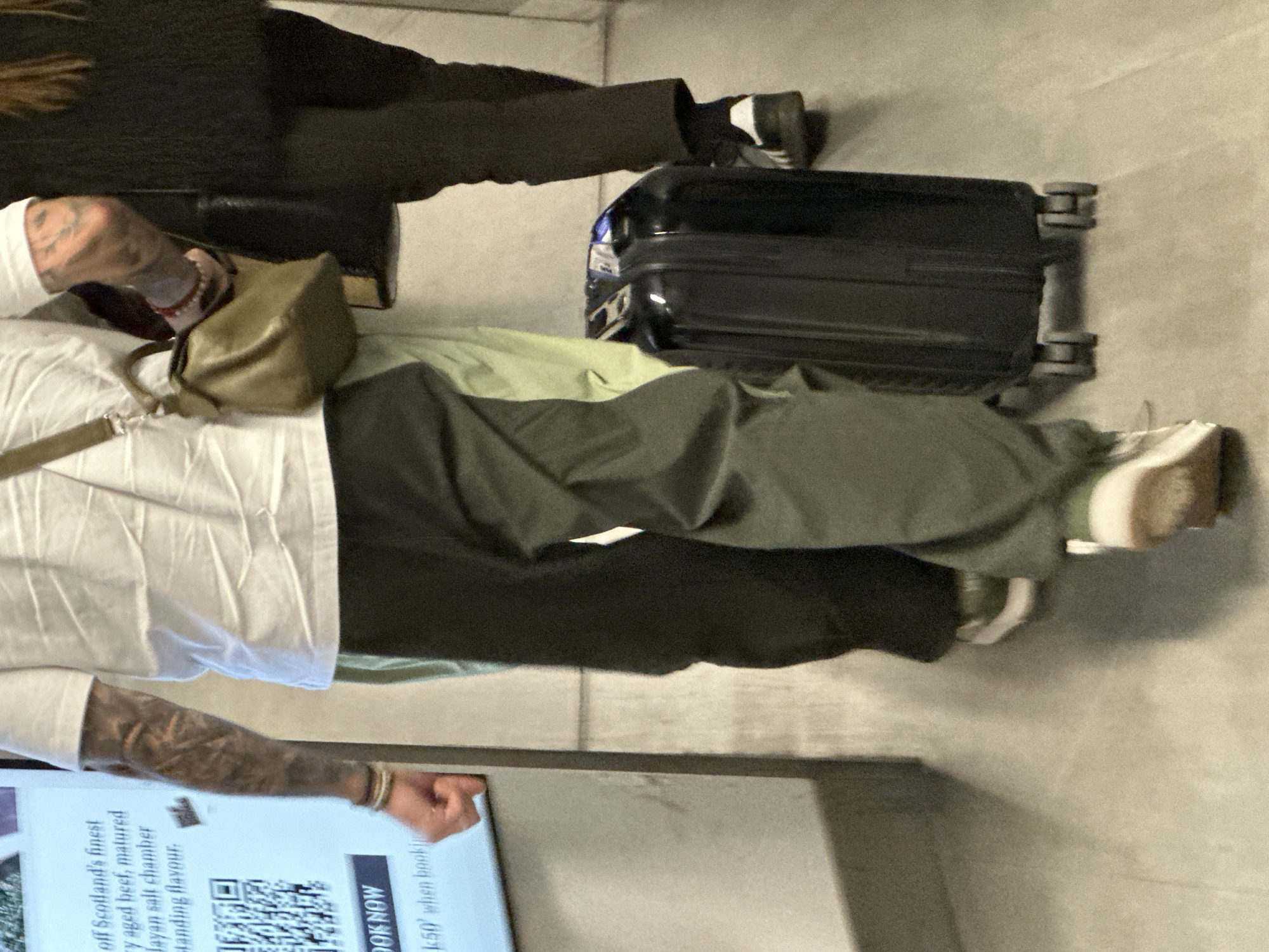 The image shows two individuals in an indoor setting, likely a public transportation terminal or similar space in the city of London, United Kingdom, as suggested by the Scottish advertising text displayed.
One individual, wearing black shoes, and black pants, is standing next to a piece of luggage, which appears to be next to an individual with multiple tattoos on their arm, wearing a white shirt, carrying a tan colored bag and olive-green pants, next to another pair of white shoes. They are in close proximity, as indicated by their luggage positions. The environment seems to be a commercial space as indicated by the advertisement on the side of the image.