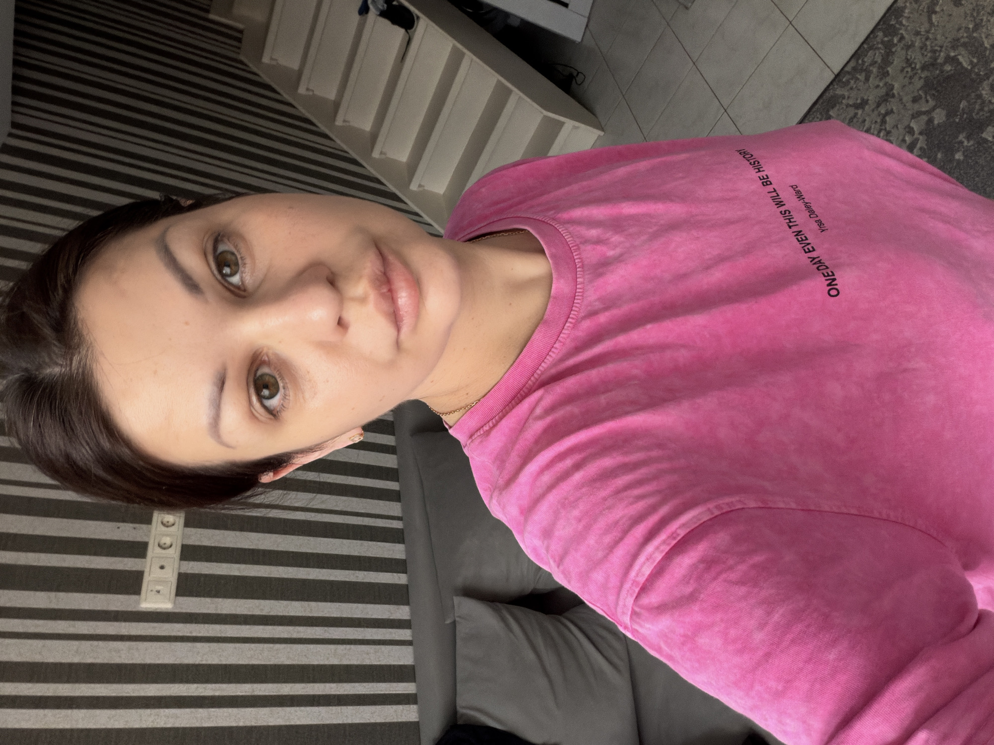 In this selfie taken indoors, a woman with dark hair and green eyes looks directly at the camera. She is wearing a pink t-shirt with text printed on it that reads, "ONE DAY EVEN THIS WILL BE HISTORY Yrsa Daley-Ward." The background features a grey couch with pillows on the left, and a white staircase on the right. The walls are striped in black and grey, and a power outlet is visible on the wall behind the woman's head. The floor is tiled. The overall impression is that of a casual indoor setting. The image does not provide any information about the city of Sochi, Russia.