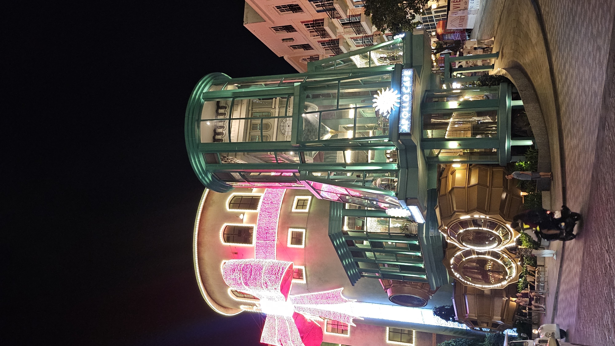 This nighttime exterior shot captures a vibrant and uniquely designed building on Phu Quoc, Vietnam, likely a part of the Grand World amusement park. The building features a whimsical, slightly steampunk aesthetic. On the left, a cylindrical structure is wrapped in bright pink lights, resembling a giant gift bow. Adjacent to it, a multi-level structure with a dark green frame and extensive glass panels houses what appears to be a cafe, indicated by the sign "APOLLO CAFE." The lower part of this building has large, circular, golden metallic elements that resemble engines or gears. The pavement in front of the building is made of cobblestones. Several people are present: a couple sits at tables outside, two individuals are on a motorcycle, and a man stands near the cafe entrance. The background shows another building with balconies and windows, illuminated and suggesting a commercial or residential area. The overall atmosphere is festive and inviting, with extensive use of colorful lights to create a captivating nocturnal scene.