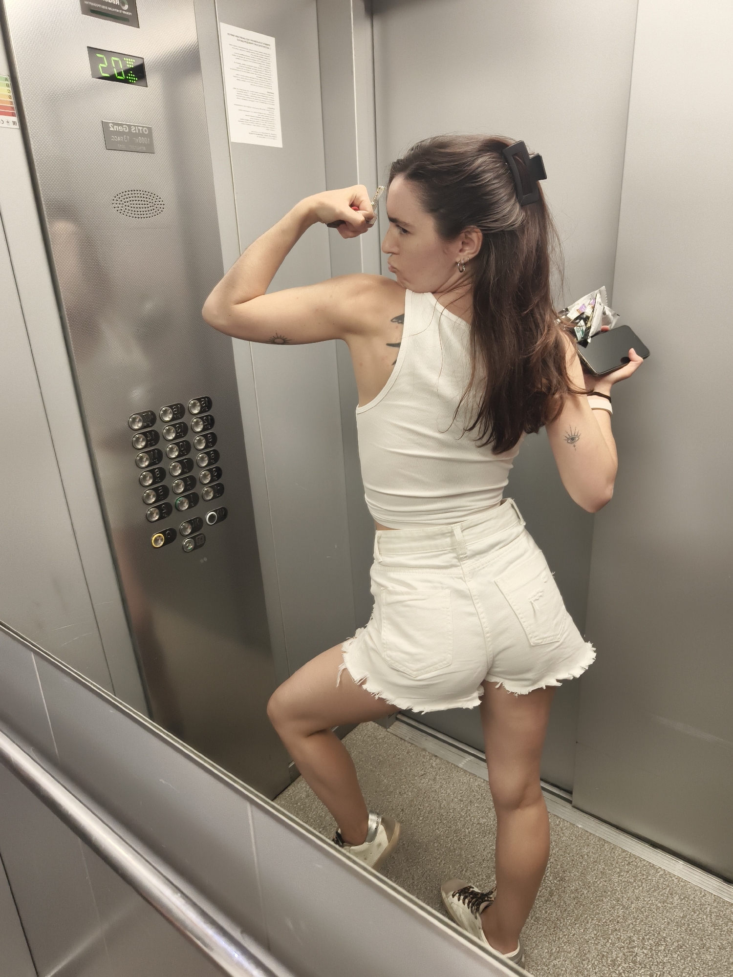 A young woman with long, dark brown hair, partially tied back with a black claw clip, poses playfully inside a modern elevator. She is captured in profile, flexing her right bicep and looking to her left with a determined yet comical expression. She is dressed in casual attire, featuring a white ribbed tank top, white distressed denim shorts with frayed edges, and white sneakers with brown laces and silver accents. Visible tattoos include a sun-like design on her right forearm/bicep and an eye-like symbol on her left forearm. She holds a black smartphone in her left hand and some small, indistinct items in her right.
The elevator interior features brushed metal walls, a grey speckled carpeted floor, and a metal handrail. On the left wall, a control panel displays numerous circular buttons for various floors, with one button illuminated in yellow. Above the buttons, a digital display shows "20" in green, indicating the elevator's current floor or destination. The brand "OTIS" and "1000R" are visible in reverse on the panel. A printed document is affixed to the back wall. This scene takes place in an elevator in Tyumen, Russia.