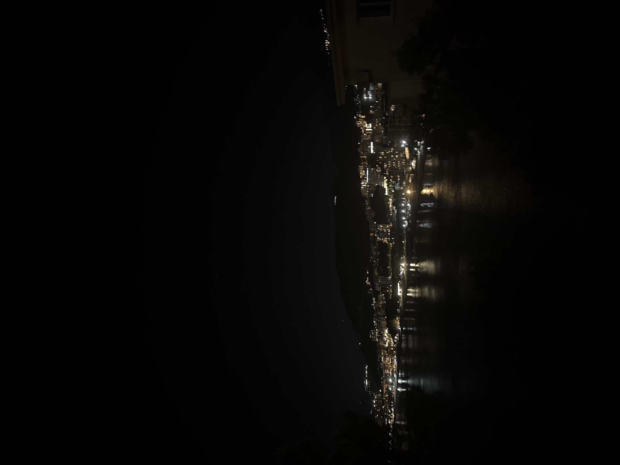 The image depicts a nighttime view of Rafailovići, Montenegro, along the coastline. The town is illuminated with numerous lights, reflecting off the water. To the left, a dark, unlit landscape or mountain range rises, creating a stark contrast. A building and trees are visible near the top right corner.