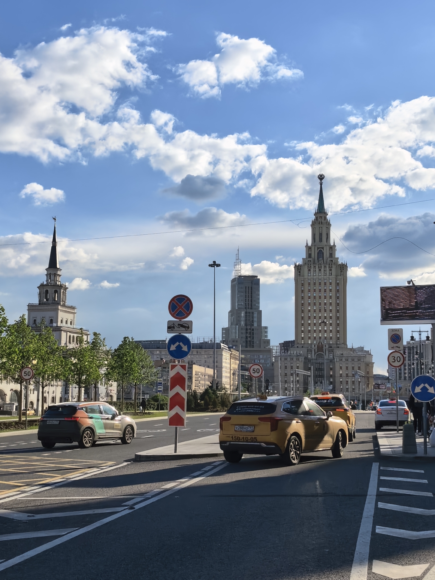 The image depicts a street scene in Moscow, Russia, with a prominent view of the Seven Sisters skyscrapers. Two are clearly visible: the Kotelnicheskaya Embankment Building, and one other. Traffic includes a silver car with green and tan accents, and yellow taxis. Various road signs are present, including speed limit and lane direction indicators. The sky features cumulus clouds.