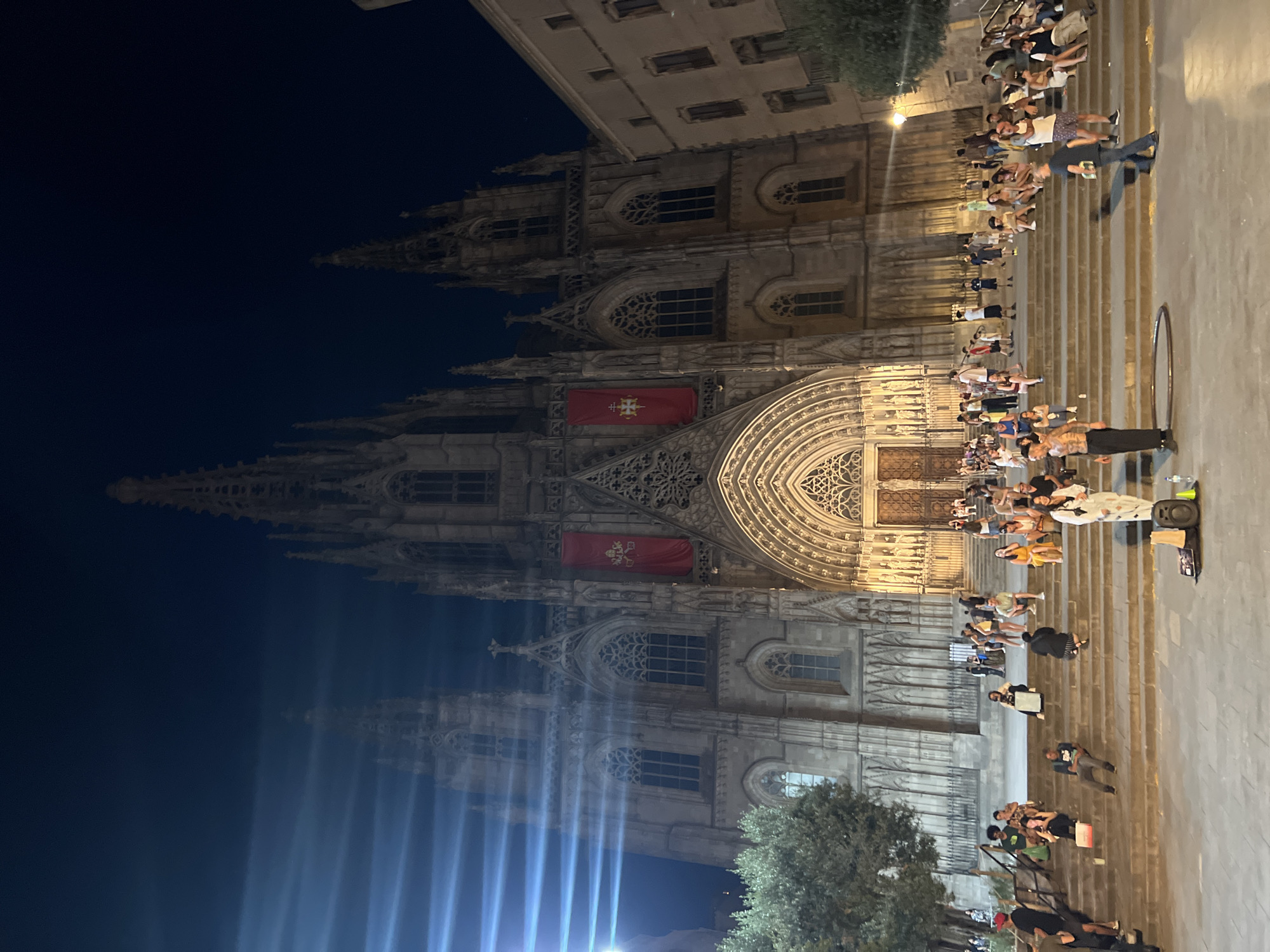 The image shows a night view of the Barcelona Cathedral in Spain. The imposing Gothic cathedral features intricate stone carvings, tall spires, and large arched entrances, all illuminated by artificial lighting. Red banners with a crest are hung on either side of the main entrance. A crowd of people is gathered on the wide, stone steps leading up to the cathedral, with some sitting, some standing, and others walking. The atmosphere appears lively, with a street performer visible in the foreground on the steps, seemingly entertaining a group. Trees and other buildings are visible to the sides of the cathedral. The sky is dark, indicating nighttime.