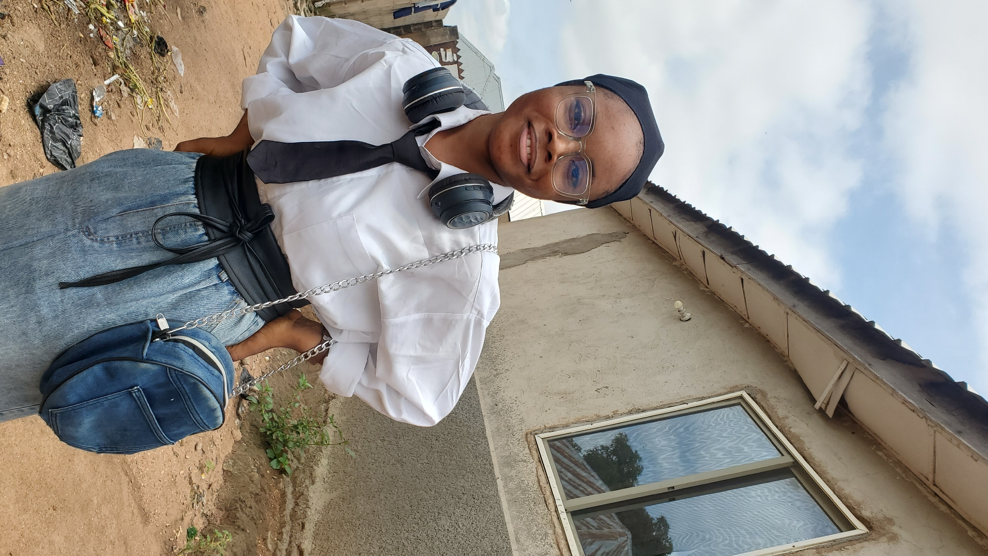 A young Black woman wearing a white button-down shirt, black tie, black headscarf, blue jeans, and clear-rimmed glasses stands with her hands on her hips outdoors. She has headphones around her neck and a denim crossbody bag with a silver chain strap. Behind her, a simple concrete building with a corrugated metal roof and a window is visible. The ground is dirt, with some debris and vegetation. The sky is overcast with clouds. The location is stated to be Jalingo, Nigeria.