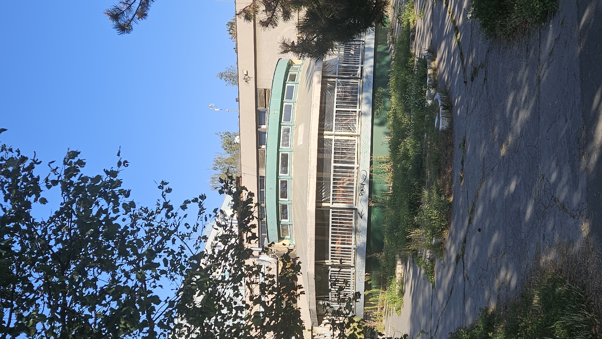 The image shows a circular building with a green trim and a light-colored awning, possibly a restaurant or cafe, at ul. Armádneho generála Svobodu in Prešov, Slovakia. The building appears somewhat weathered, with overgrown vegetation surrounding it and graffiti on its lower section. The area in front of the building is paved with cracked asphalt and interspersed with patches of weeds and grass. A tree with dark green foliage partially obscures the view of the building from the left side of the frame. The sky is clear and blue. No people are visible in the image.