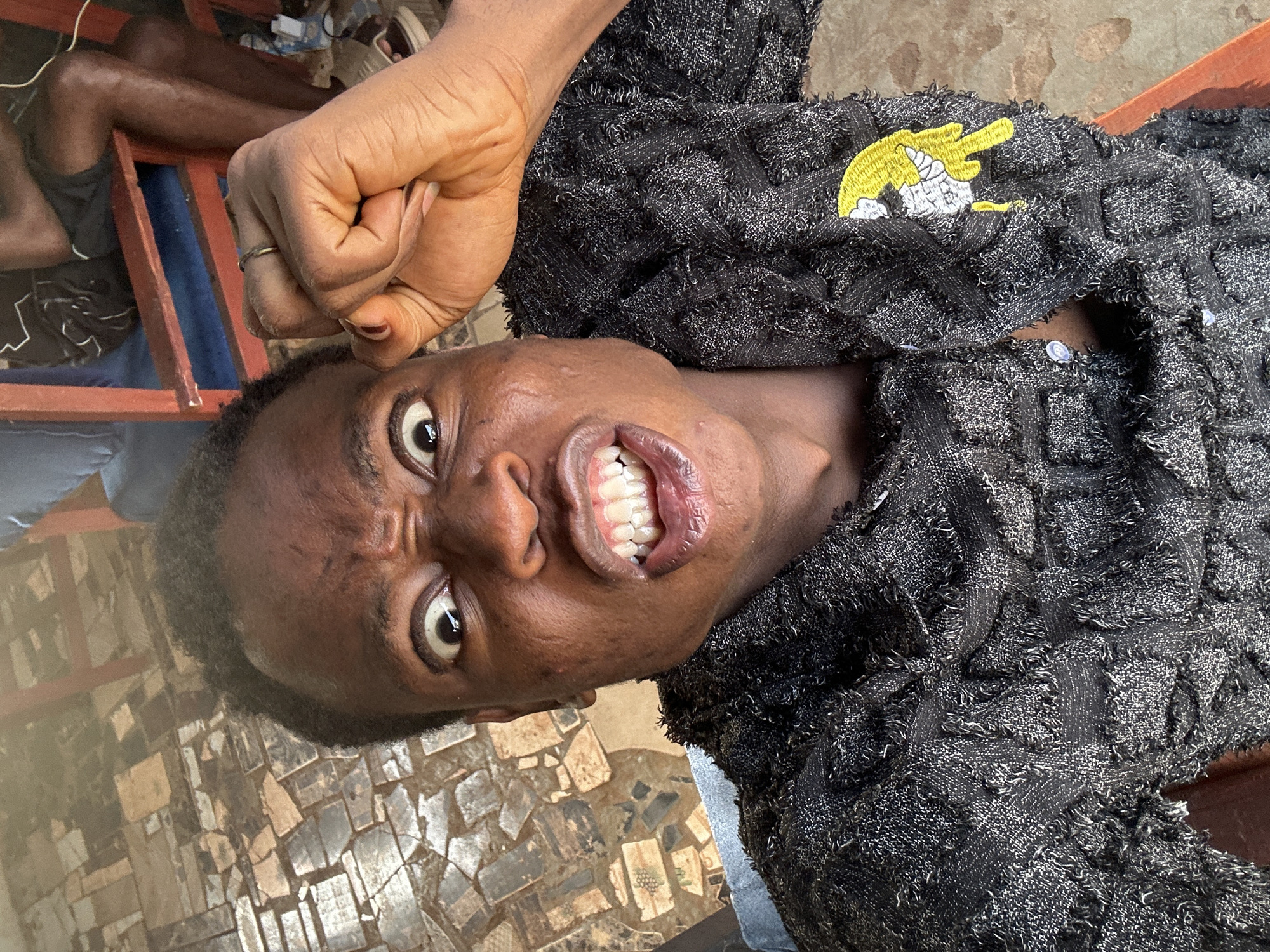 A close-up shot captures a person with dark skin and short, dark hair. The person is wearing a black, textured jacket with a yellow embroidered patch on the right side. Their face is contorted in an expression of anger or strain, with furrowed brows, wide eyes, and clenched teeth. The person's right hand is clenched into a fist and raised.
The person appears to be seated outdoors. The background consists of a mosaic-patterned ground and the legs of other people seated nearby, as well as furniture elements.