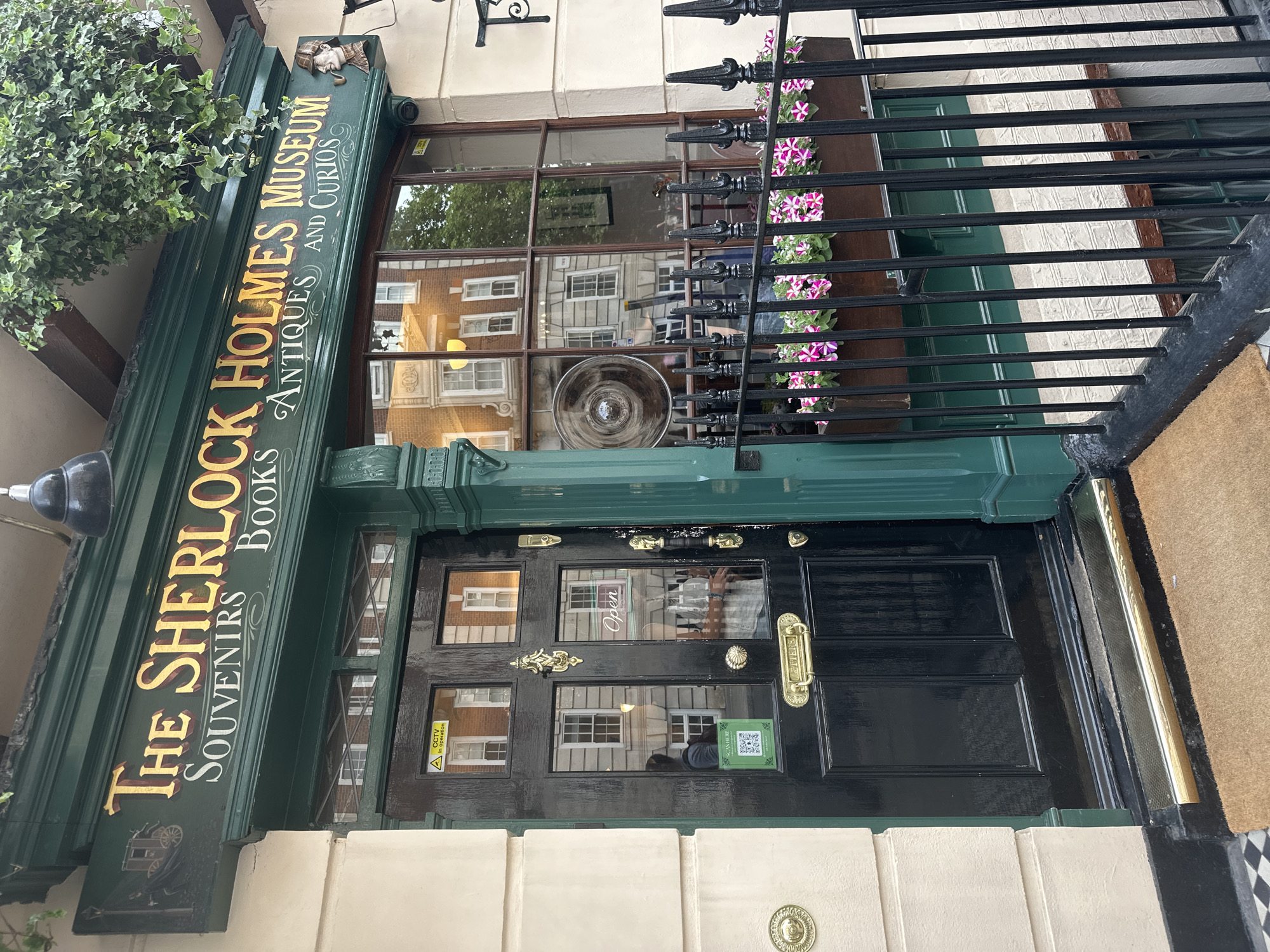 The image depicts the entrance to The Sherlock Holmes Museum, located in London, United Kingdom. The museum's sign, painted in dark green with gold lettering, reads "The Sherlock Holmes Museum, Souvenirs, Books, Antiques and Curios." The entrance is framed by a black iron fence with flower boxes containing pink and white flowers. The black door has four small glass windows and ornate brass hardware. A small sign with a QR code is visible on the door, as well as a CCTV sign. The building facade is light-colored, with cream-colored stone or stucco on the lower section. The reflection in the door and window shows buildings across the street. A doormat is partially visible on the right side of the frame.