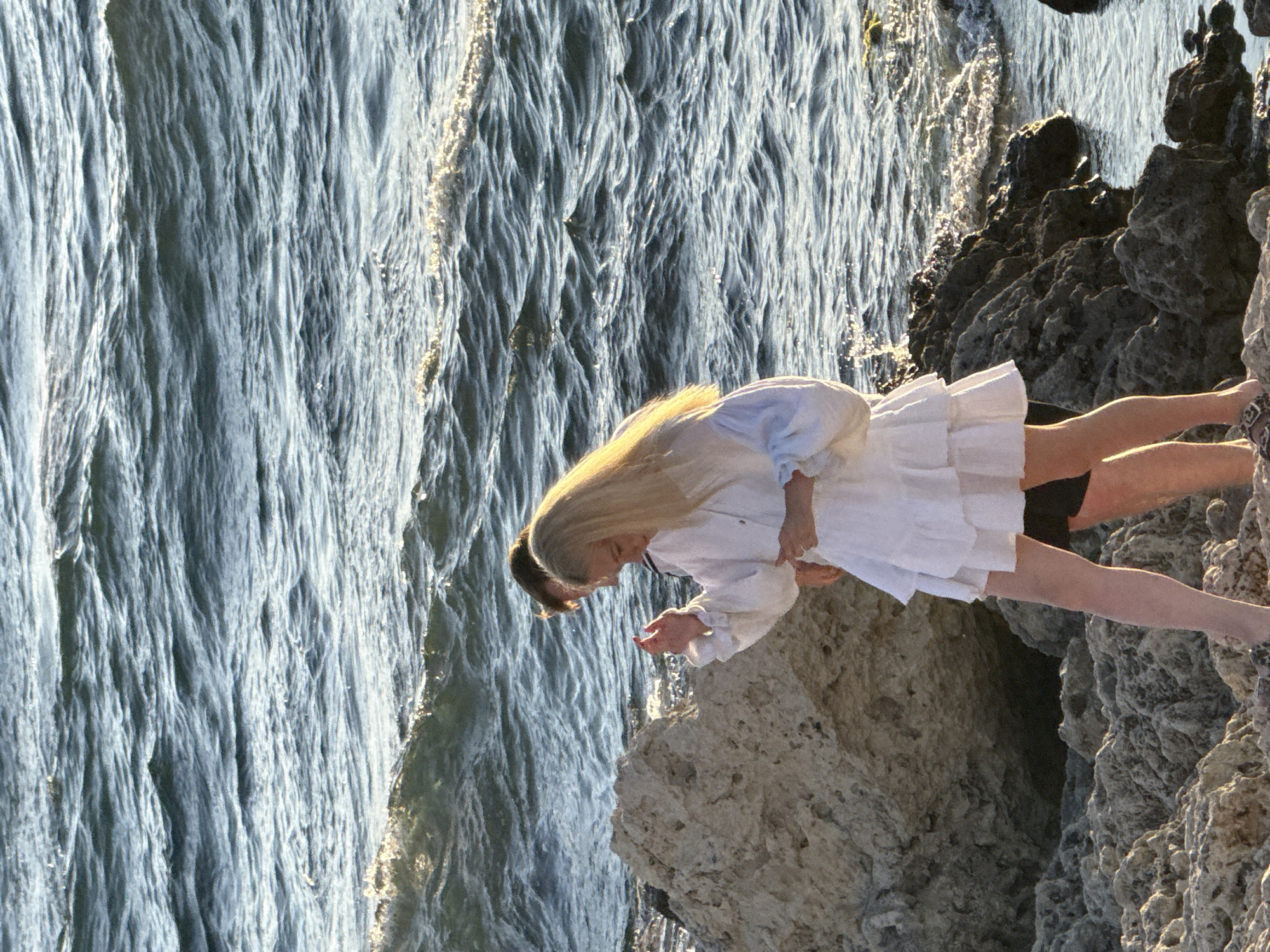 This media file shows two people near a rocky coastline with the ocean in the background. One person, with long blonde hair, is wearing a white ruffled dress and appears to be leaning over the rocks. The other person, mostly out of frame, has dark hair and is also near the edge. The water is choppy and reflects the sunlight, suggesting it is daytime, possibly late afternoon given the warm light. The overall scene evokes a sense of peacefulness and perhaps a touch of adventure as they are close to the edge of the water. There is no visible text in the image.