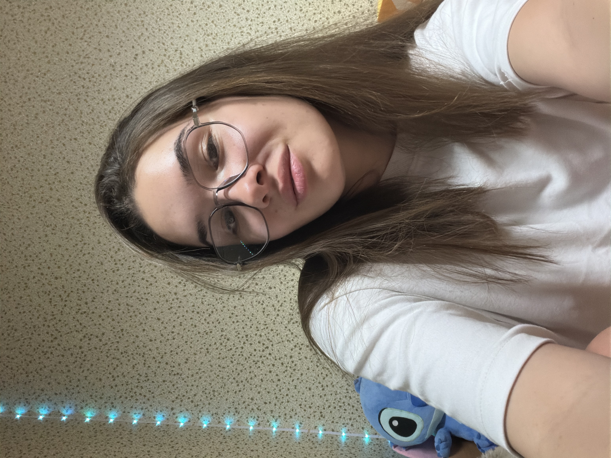 A selfie taken at an angle. The subject is a young woman with brown hair and glasses, wearing a white t-shirt. A blue plush toy, possibly "Stitch", is visible near her arm. A string of small, blue, decorative lights is in the lower portion of the frame. The background appears to be a textured wall. There is no discernible information in the image to suggest the location is Sofia, Bulgaria.
