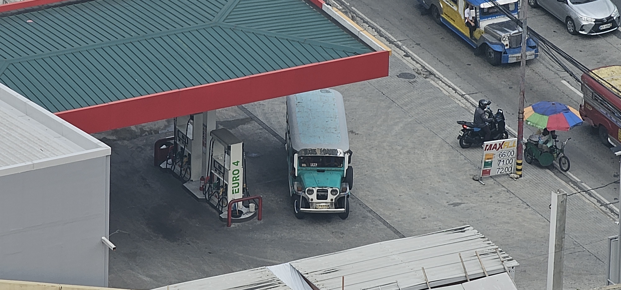 The image depicts a scene at a gasoline station in Makati, Philippines. A green and teal jeepney is parked next to a gas pump with "EURO 4" clearly visible on the pump. To the right, a motorcycle rider is seen heading towards a sign displaying fuel prices: 66.00, 71.00, and 72.00. Further to the right, a vendor is operating under a colorful umbrella, with a food cart. In the background, a silver car and a yellow and blue jeepney are visible on the road. The environment appears to be an urban street scene, with buildings and overhead wires present.