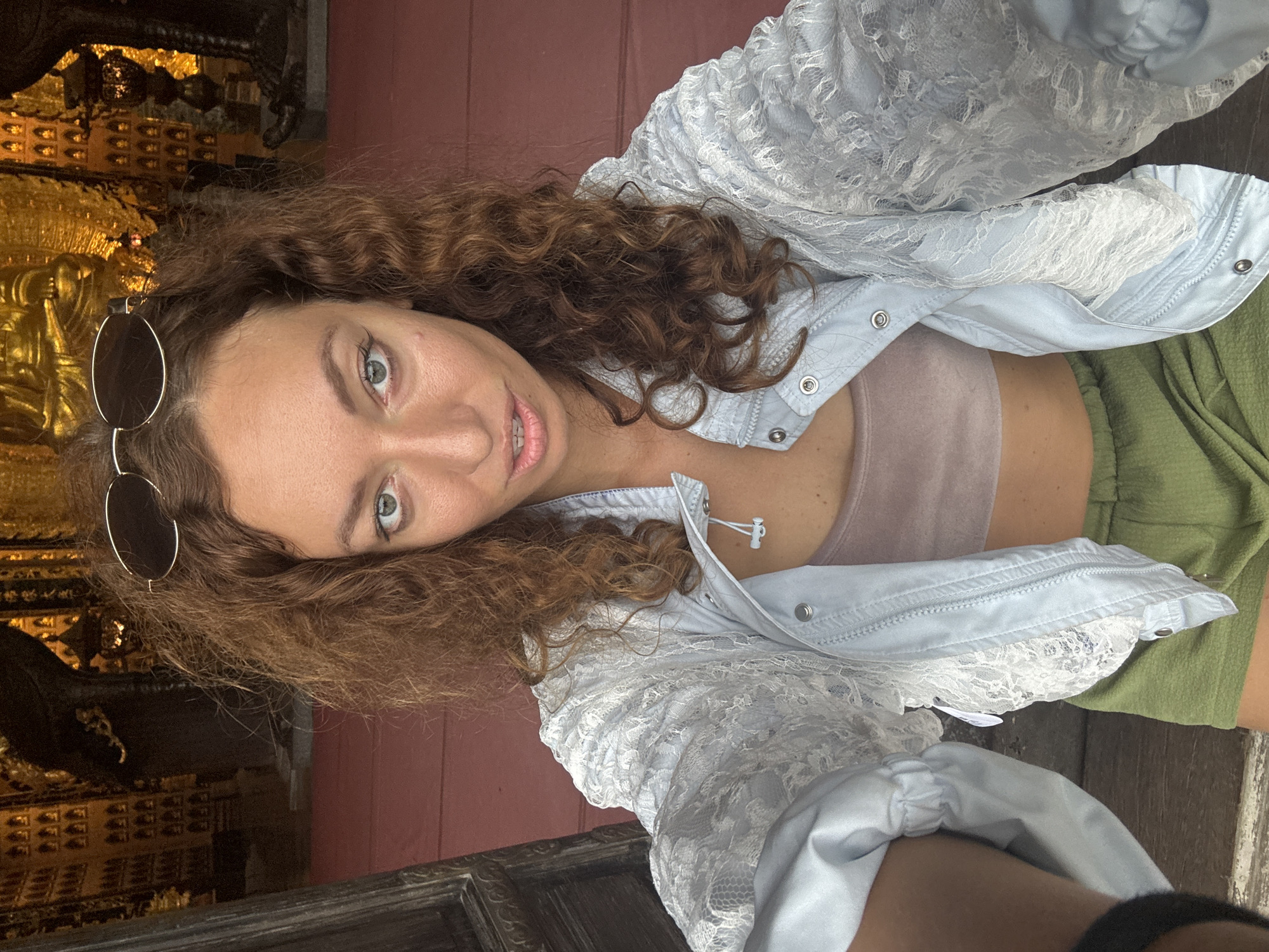A woman with curly brown hair and green eyes is taking a selfie in what appears to be a temple or religious building in Tây Hoa Lư Ward, Vietnam. She is wearing a light blue lace jacket over a pink crop top and green shorts. Sunglasses are perched on top of her head. The background is ornate and golden, with rows of small Buddha statues and larger statues visible. The floor is covered in a red carpet. The overall lighting is warm and inviting.