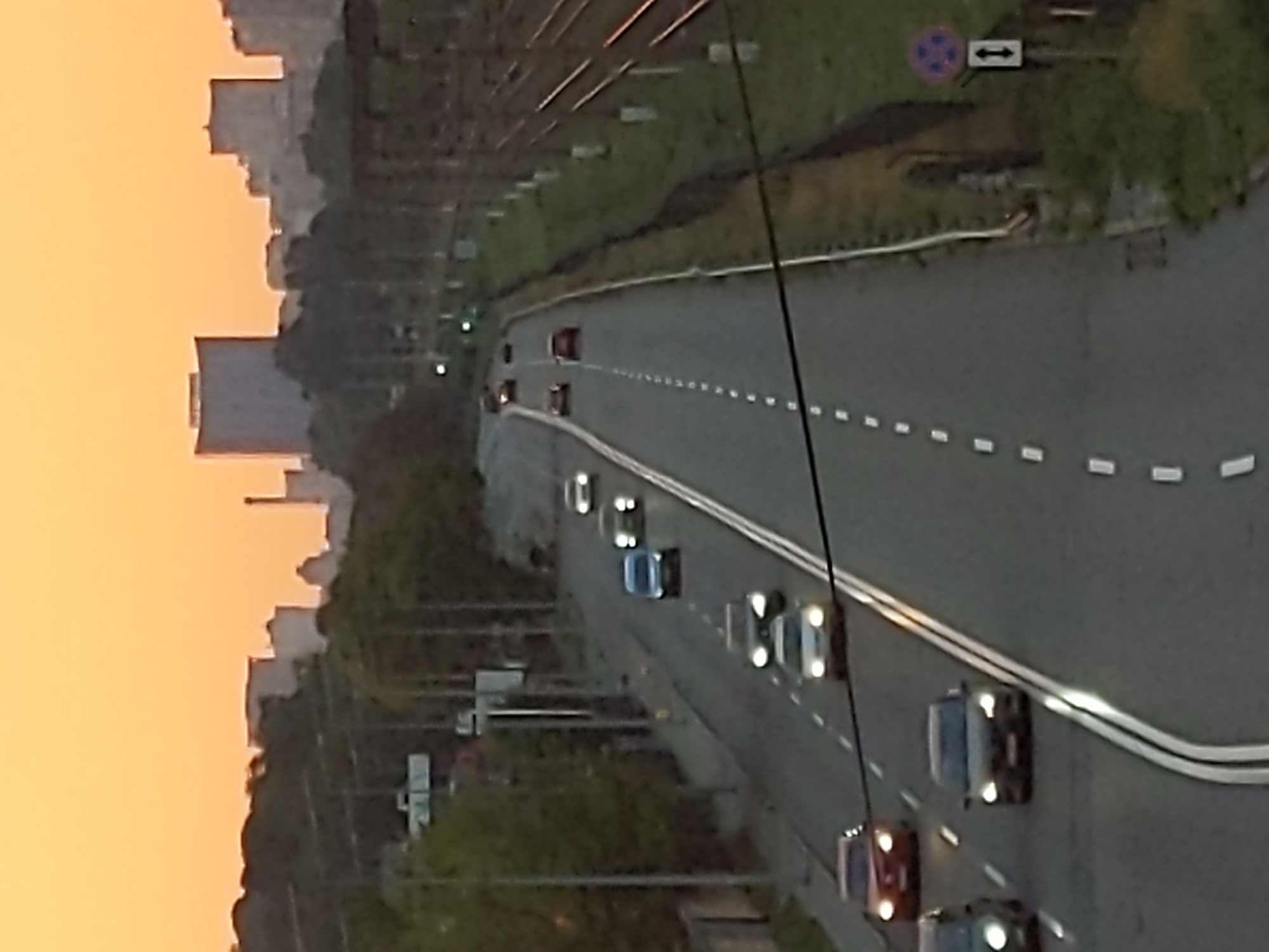 The image shows a view of the Kharkiv Highway in Kyiv, Ukraine, during what appears to be sunset or dawn, indicated by the warm orange sky. Multiple cars are driving on the multi-lane asphalt road, which has white dashed lane markers. On the right side of the road, there are railway tracks and a grassy embankment. The background features a city skyline with several buildings, including what looks like a modern high-rise and some older structures. Trees line the left side of the highway. A traffic sign with a symbol of a red circle with four blue dots and an arrow pointing up and down is visible on the right.