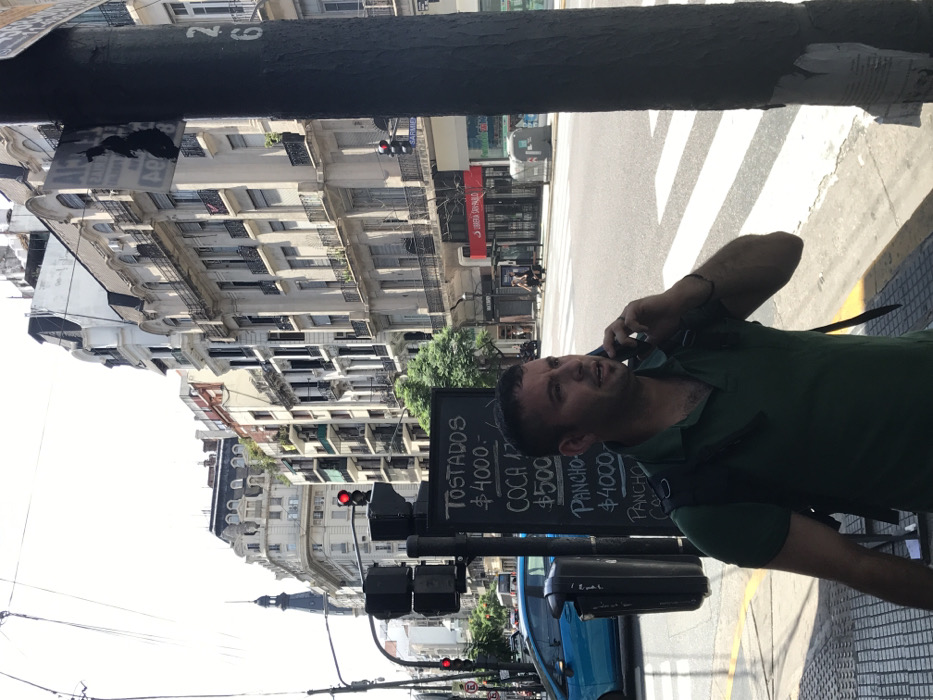 A man in a green shirt talks on his cell phone on a city street. A menu board advertises "Tostadas $4000," "Coca $500," and "Pancho $4000." Buildings with balconies line the street, and a crosswalk is visible. Traffic lights hang overhead. A blue car is parked nearby. A black pole is in the foreground.