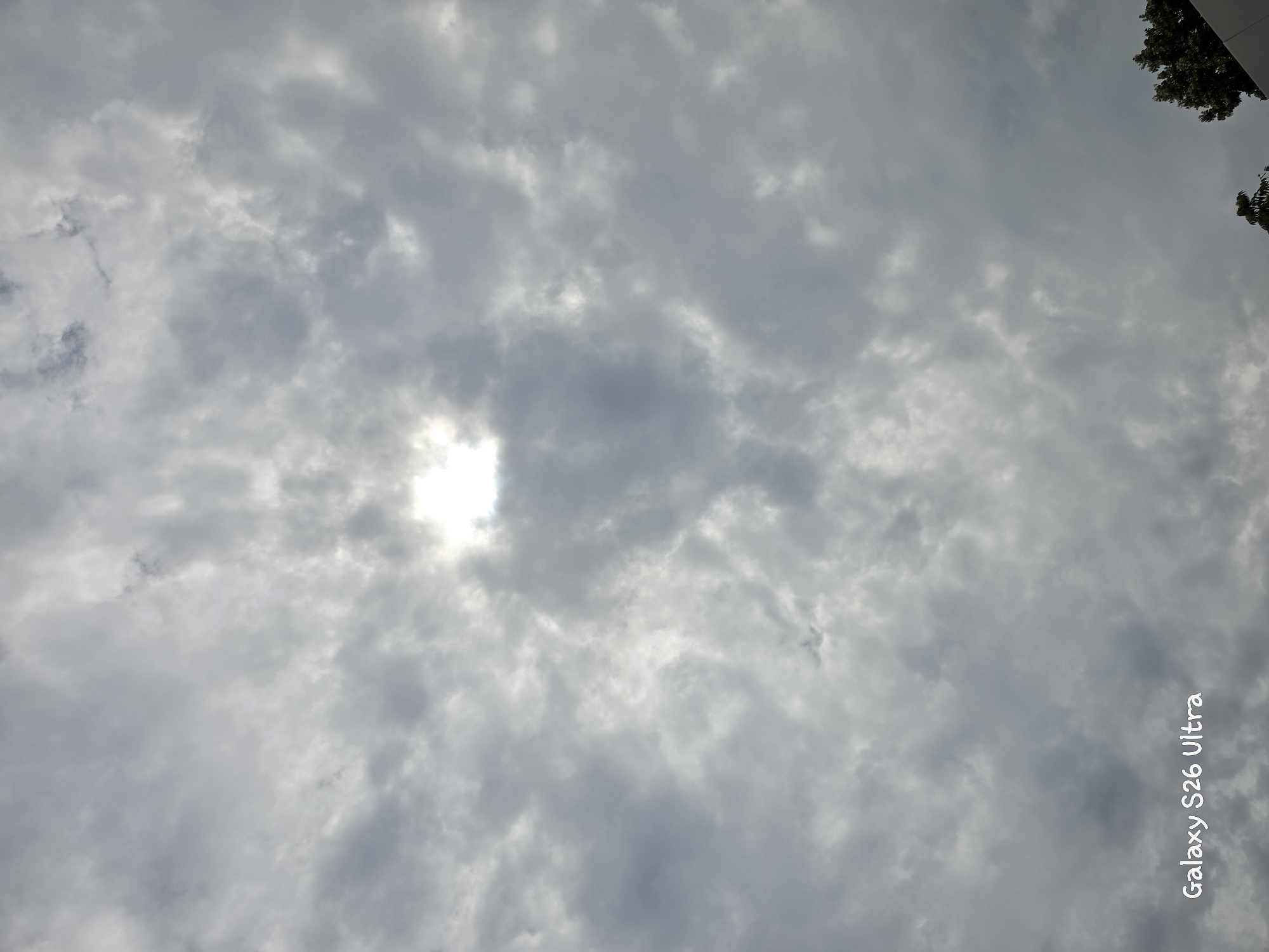 The image depicts a cloudy sky with the sun peeking through a break in the clouds. The sky is overcast with various shades of gray clouds, suggesting a partly cloudy or overcast day. The sun appears as a bright, diffused light source at the top center of the frame. In the lower left corner of the image, the text "Galaxy S26 Ultra" is visible, indicating the device used to capture the photo and a potential timestamp or location tag. In the lower right corner, the top of a tree with dark green foliage and a sliver of a white structure, possibly a building, are visible. The overall scene is a natural outdoor environment, likely taken from ground level looking upwards. There are no people or specific Plovdiv landmarks identifiable in this image.