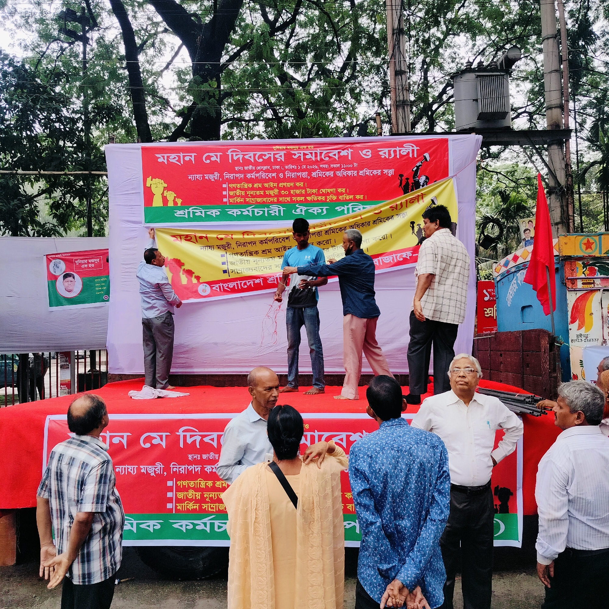 An outdoor scene captures a public gathering or rally, likely in Bangladesh. A temporary stage, covered in red fabric, serves as the focal point, with several men on it. One man in a light blue shirt adjusts a banner, while a young man in a blue and white t-shirt stands barefoot with an older man, who holds a red string or rope, seemingly engaged in a demonstration or activity. A crowd of men, and one woman seen from behind, are gathered on the ground, observing the stage or conversing.
Large banners, predominantly red and yellow, decorate the stage and surrounding area, displaying text in Bengali. The main banner announces a "Great May Day Gathering and Rally," specifying the location as "National Press Club, Dhaka," and the date as "May 1, 2026, at 11:00 AM." It lists demands for "Fair wages, safe working environment and security, worker's rights, worker's identity," followed by bullet points: "Enact democratic labor law," "Declare national minimum wage 30 thousand taka," and "Cancel all harmful agreements with American imperialism." The organizing body is identified as the "Workers-Employees Unity Council." Another banner on the left reads "Workers of the world unite" and mentions "BNP," accompanied by a portrait.
The background features lush green trees, suggesting an outdoor urban setting. Utility poles with an electrical transformer are visible, along with a red flag and part of a blue vehicle, possibly a truck. The event appears to be an organized public demonstration or commemoration of May Day, advocating for workers' rights.
