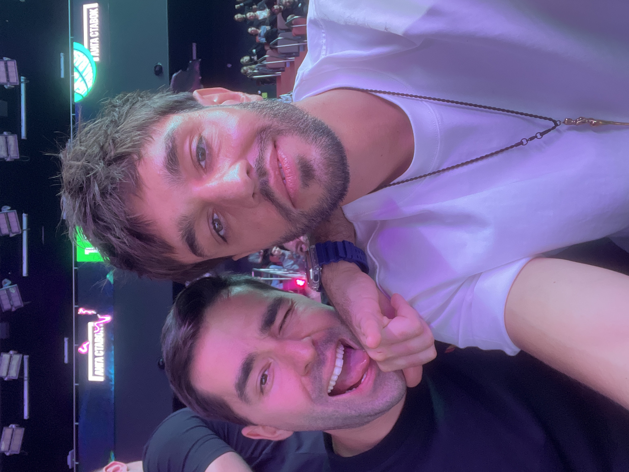 Two young men are posing for a selfie-style picture indoors. The man on top, wearing a white T-shirt and a necklace, looks directly at the camera. The man on the bottom is wearing a black T-shirt, winking, sticking his tongue out, and appears to be playfully restrained with the arm of the other man. A blue wristwatch is visible on the arm used for the playful restraint.
In the background, there are rows of seats filled with people, suggesting a theater, concert hall, or similar venue. Two illuminated signs are visible: one in green and one in pink, which include Cyrillic text. Judging by the Cyrillic text "АМТА СТАВОК" in pink letters, the location is most likely the Aktsionernoe Obshchestvo "Moskovskaya Mezhregional'naya Transportnaya Assotsiatsiya 'Tsentr," Moscow, Russia. Several stage lights hang in the background. The lighting in the room is predominantly purple.