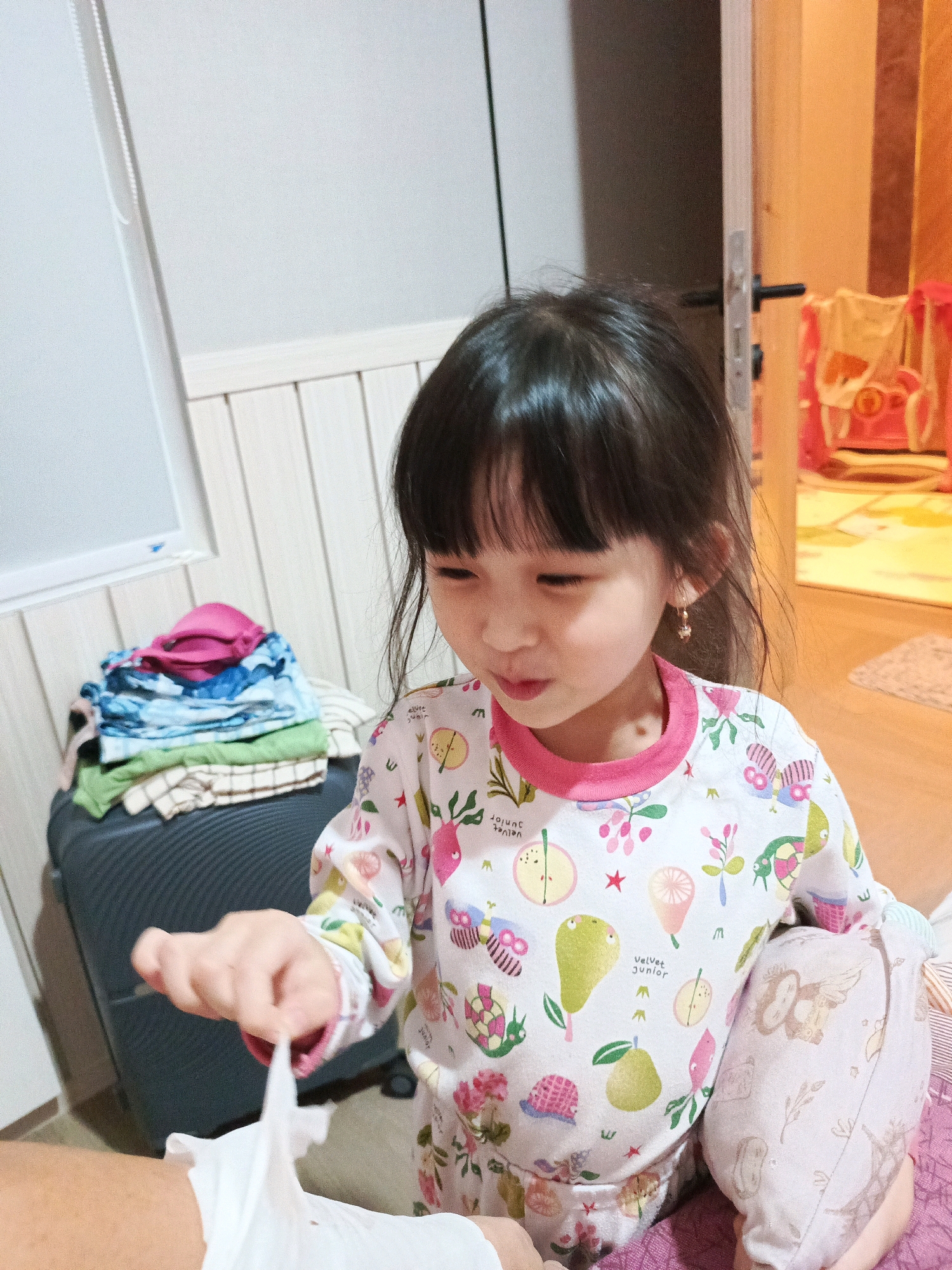 A young girl with dark hair and bangs is looking down at her hands, which are holding a white tissue. She is wearing a long-sleeved shirt with a colorful fruit and floral pattern, and pink trim around the neck. The shirt has text that appears to read "Velvet junior". She is also wearing pants with a similar print, featuring owls and the Eiffel Tower. The girl has a small gold earring in her right ear.
In the background, to the left, is a dark gray suitcase with a pile of folded clothes on top. The clothes are a mix of colors and patterns, with a bright pink item visible on top. The wall behind the suitcase appears to be paneled wood. To the right, a door is slightly ajar, revealing a brightly lit room with vertical stripes of color on the wall. A pink crib or playpen with toys is visible in the background room.
The lighting suggests it might be indoors during the day or evening. The scene appears to be in a domestic setting, possibly a bedroom or living area. The girl's facial expression is one of concentration or slight distress as she manipulates the tissue. The overall impression is a candid moment captured in a home environment.