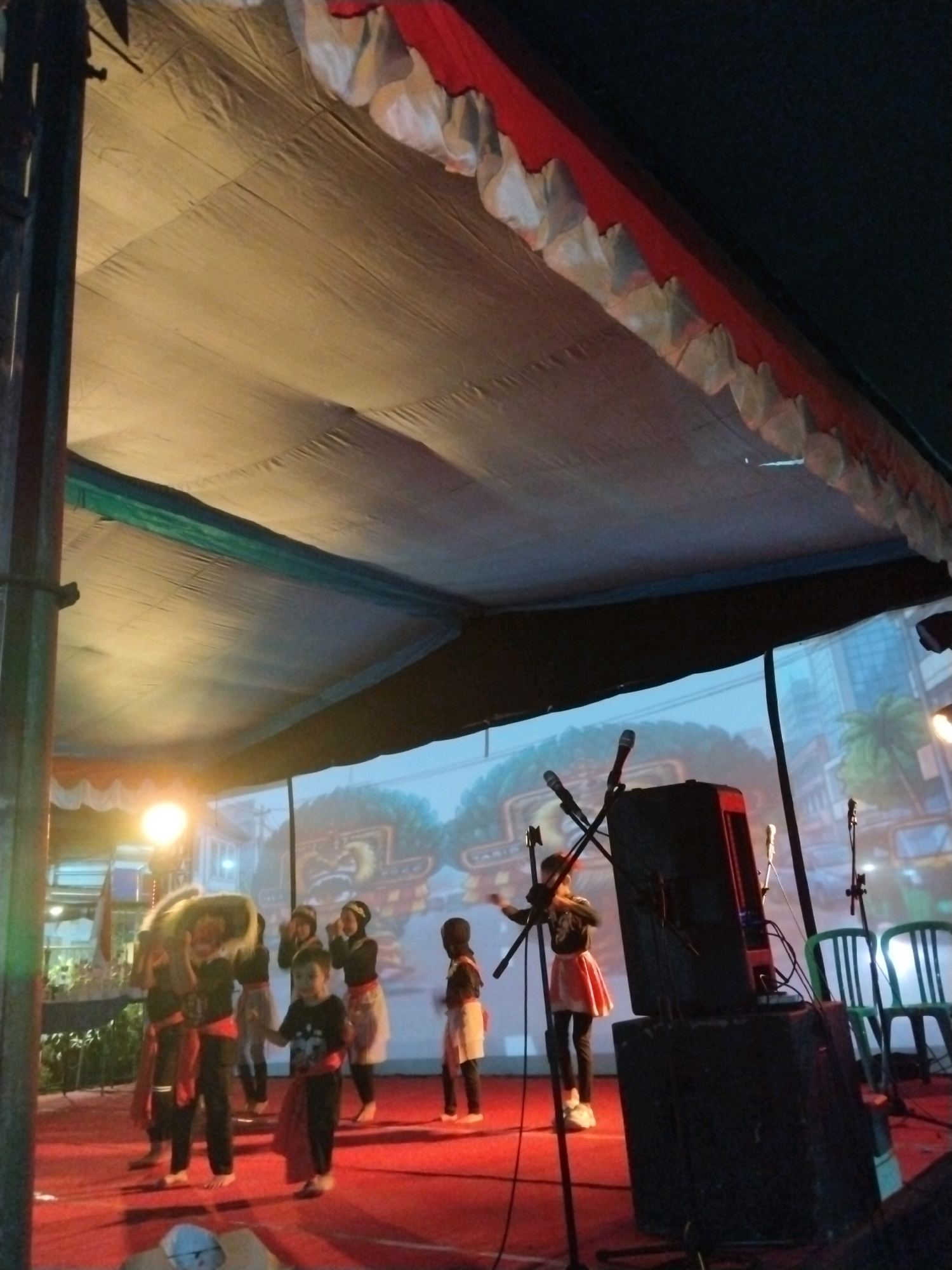 The image shows a performance on a stage in Singosari, Indonesia. A group of children, dressed in traditional attire, are performing a dance. One child in the front is holding a large, lion-like mask. The stage is covered with a red carpet. Behind the stage, there is a large screen displaying an animated cityscape. Several microphones and speakers are set up on the stage, suggesting that the performance is being amplified. The scene is illuminated by stage lights. The performance appears to be taking place outdoors, under a large tent.