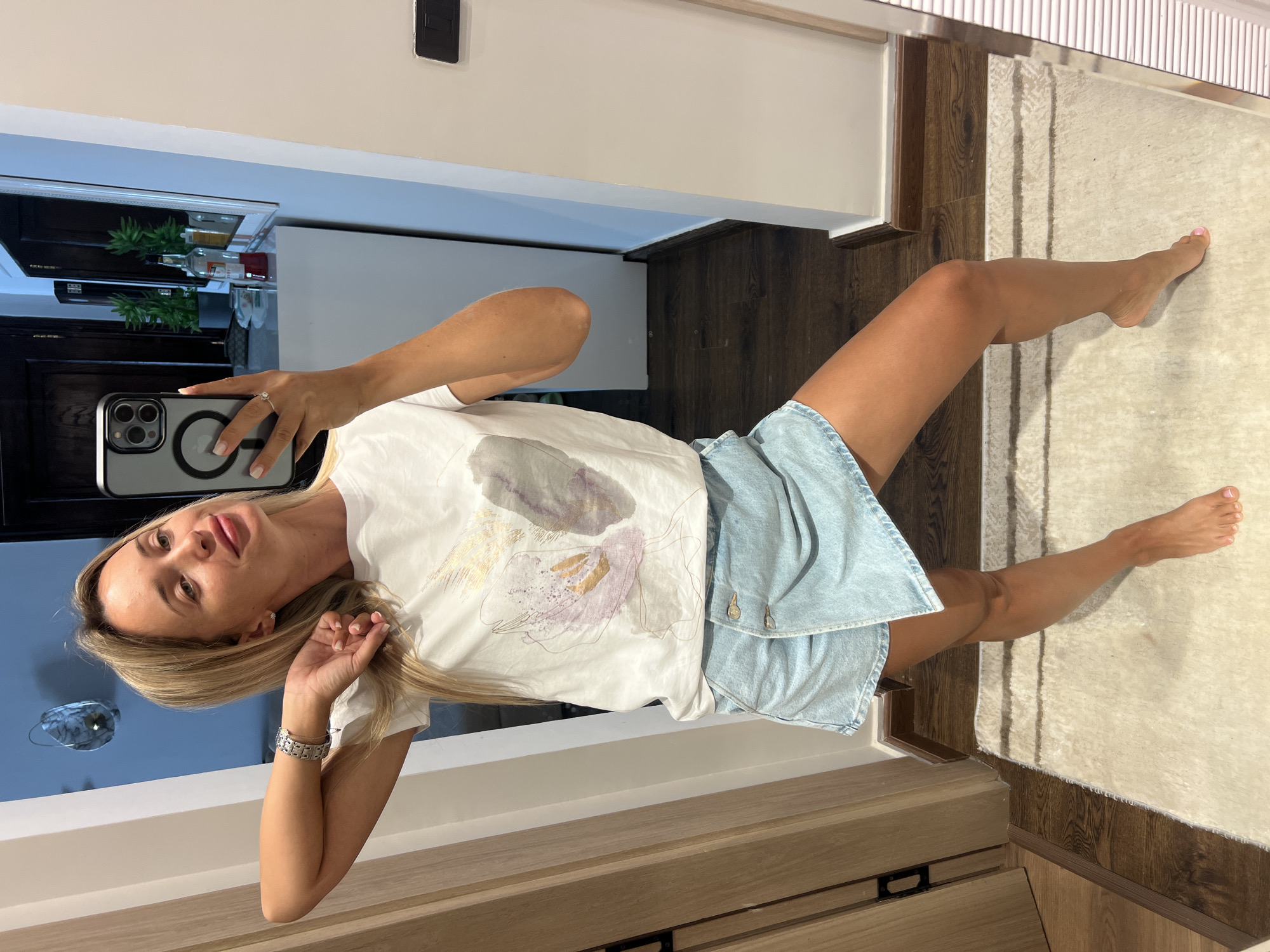A woman with blonde hair, wearing a white t-shirt with a floral print and a light blue denim skirt, is taking a selfie in a hallway with mirrored walls. She is standing on a light-colored rug, with wooden floors visible in the background. The hallway appears to be in a modern residential or hotel setting, likely within Sharm El Sheikh, Egypt. The woman is posing with her left arm bent and hand on her head, looking towards the camera. The overall impression is casual and personal, suggesting a moment of self-appreciation or documenting an outfit.