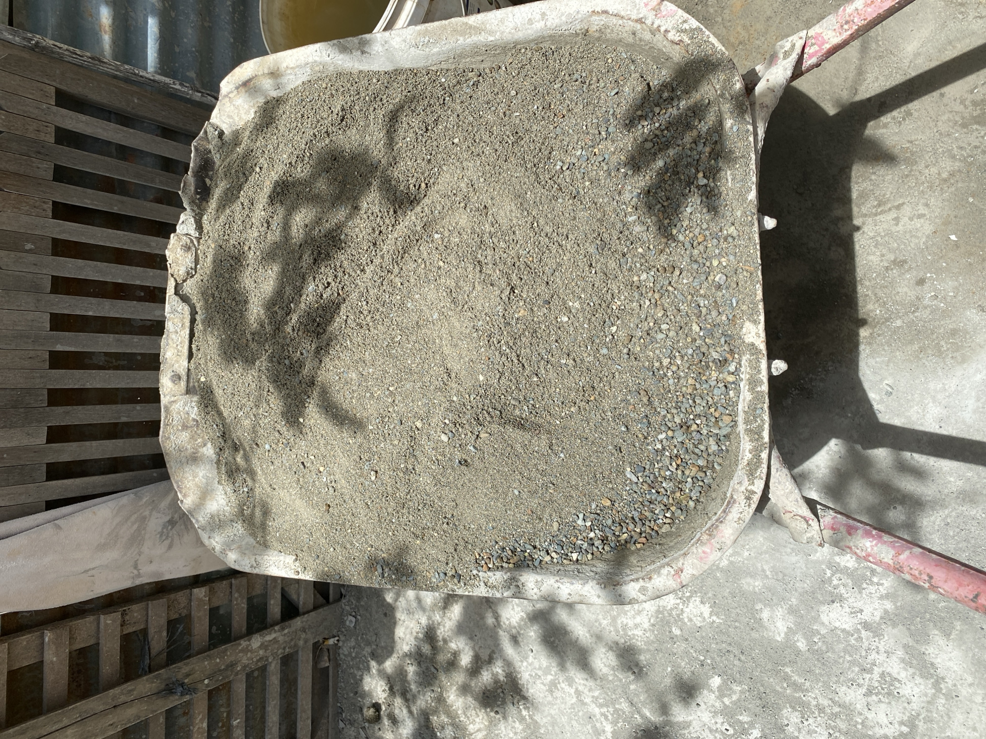 A high-angle, close-up shot shows a wheelbarrow full of sand and gravel. The wheelbarrow's basin is roughly half-full with the mixture, which is a light tan color with small grey and brown stones mixed in. The edge of the wheelbarrow is chipped and worn, indicating heavy use. In the background, wooden planks are visible, possibly part of a fence or structure. Sunlight casts dappled shadows of leaves onto the sand and the ground surrounding the wheelbarrow. The ground appears to be concrete with some scattered debris. A hint of a yellow container is visible to the right of the wheelbarrow. The scene is outdoors, likely in a construction or work area.