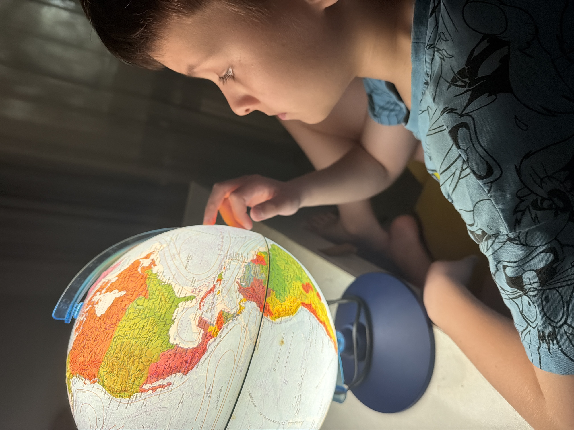A young boy is intently examining a globe. He is positioned close to the globe, with his finger resting on its surface. The globe is illuminated and displays a detailed map of the continents. The boy is wearing a blue shirt with cartoon characters printed on it. The environment appears to be indoors, possibly a room with dim lighting. There is no evidence to suggest this image was taken in the city of Votkinsk, Russia.