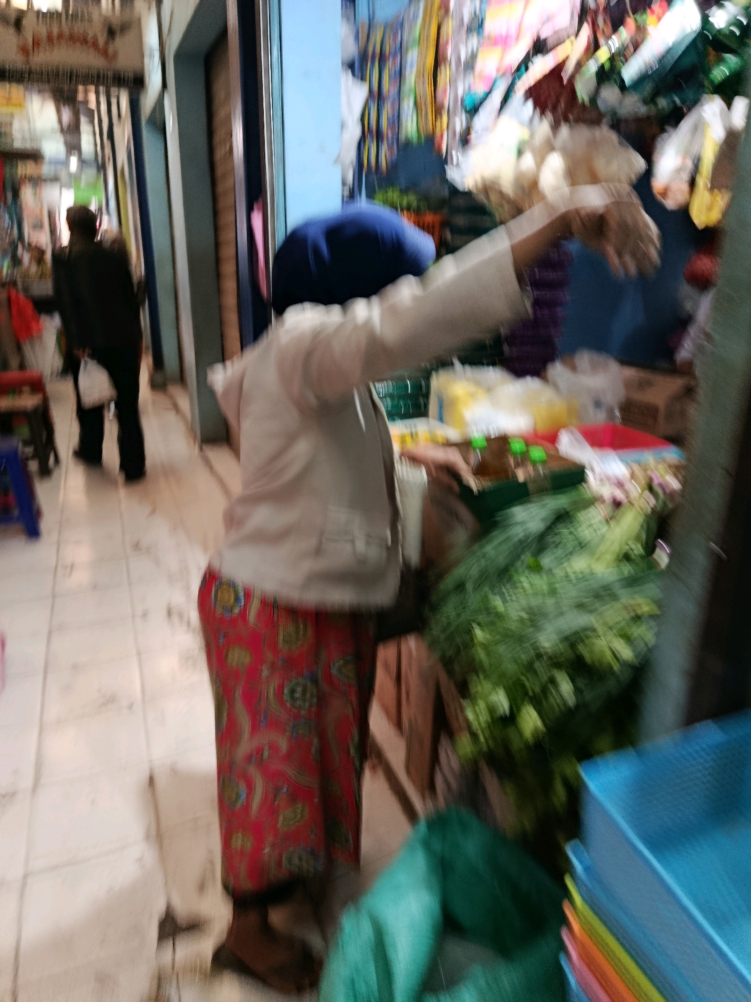The image depicts a blurred, indoor scene likely at a market in Bululawang, Indonesia. A person in the foreground, wearing a white jacket and a red patterned skirt, is standing near a stall with vegetables. The person's face is not visible. Behind them, another person in dark clothing is walking away, carrying a white plastic bag. The environment includes shelves or stalls displaying goods, possibly food items like vegetables and packaged products. The floor is tiled. The overall impression is a busy market setting, though the blurriness obscures many details.