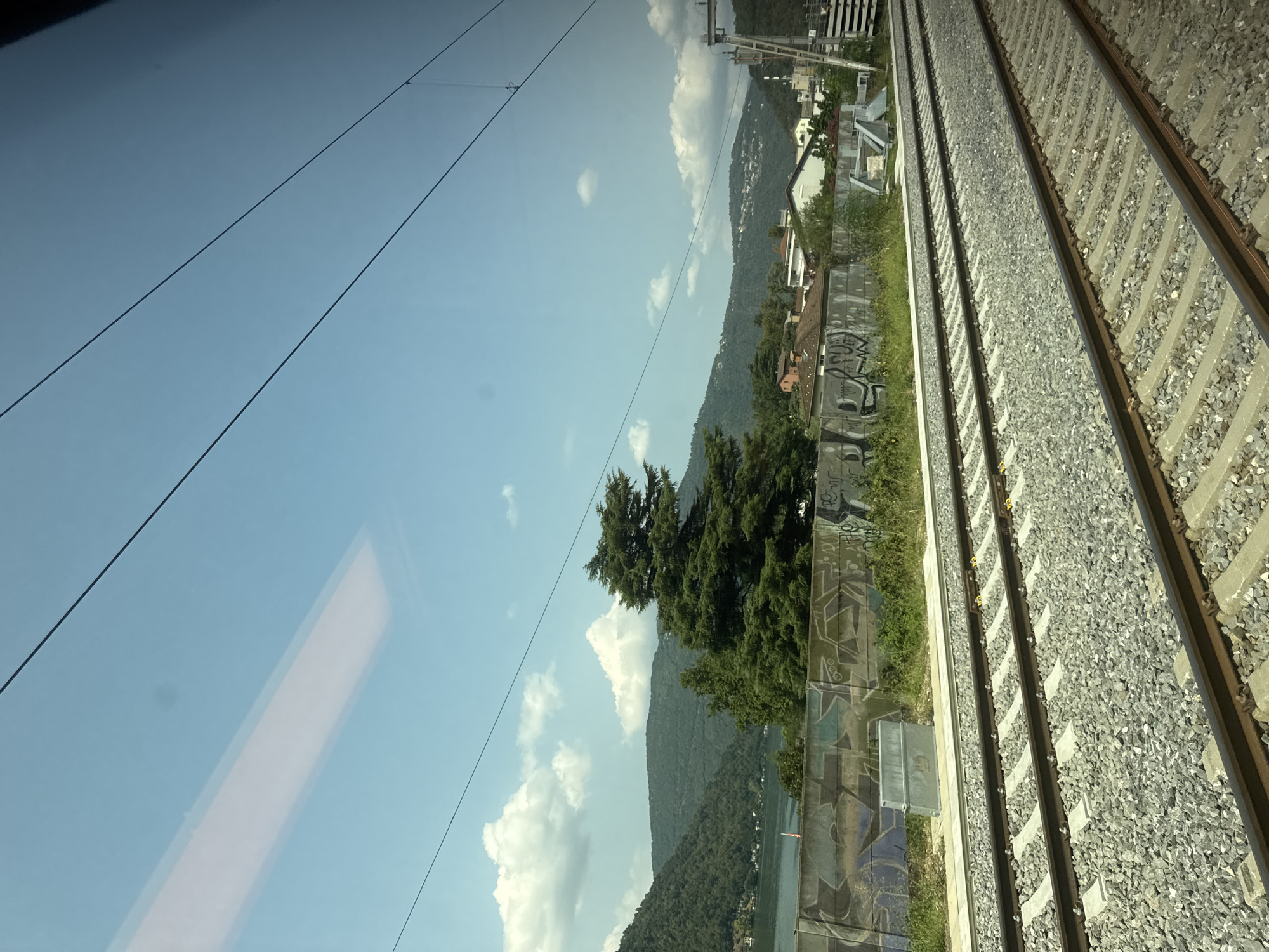 A view from a train window shows a body of water, hills covered in trees, and a town in the distance. The train tracks are in the foreground, with gravel ballast and concrete ties. A concrete wall with graffiti runs along the side of the tracks. The sky is blue with scattered white clouds. The scene is likely in Val Mara, Switzerland, given the context of the image. There are no people visible in the image.