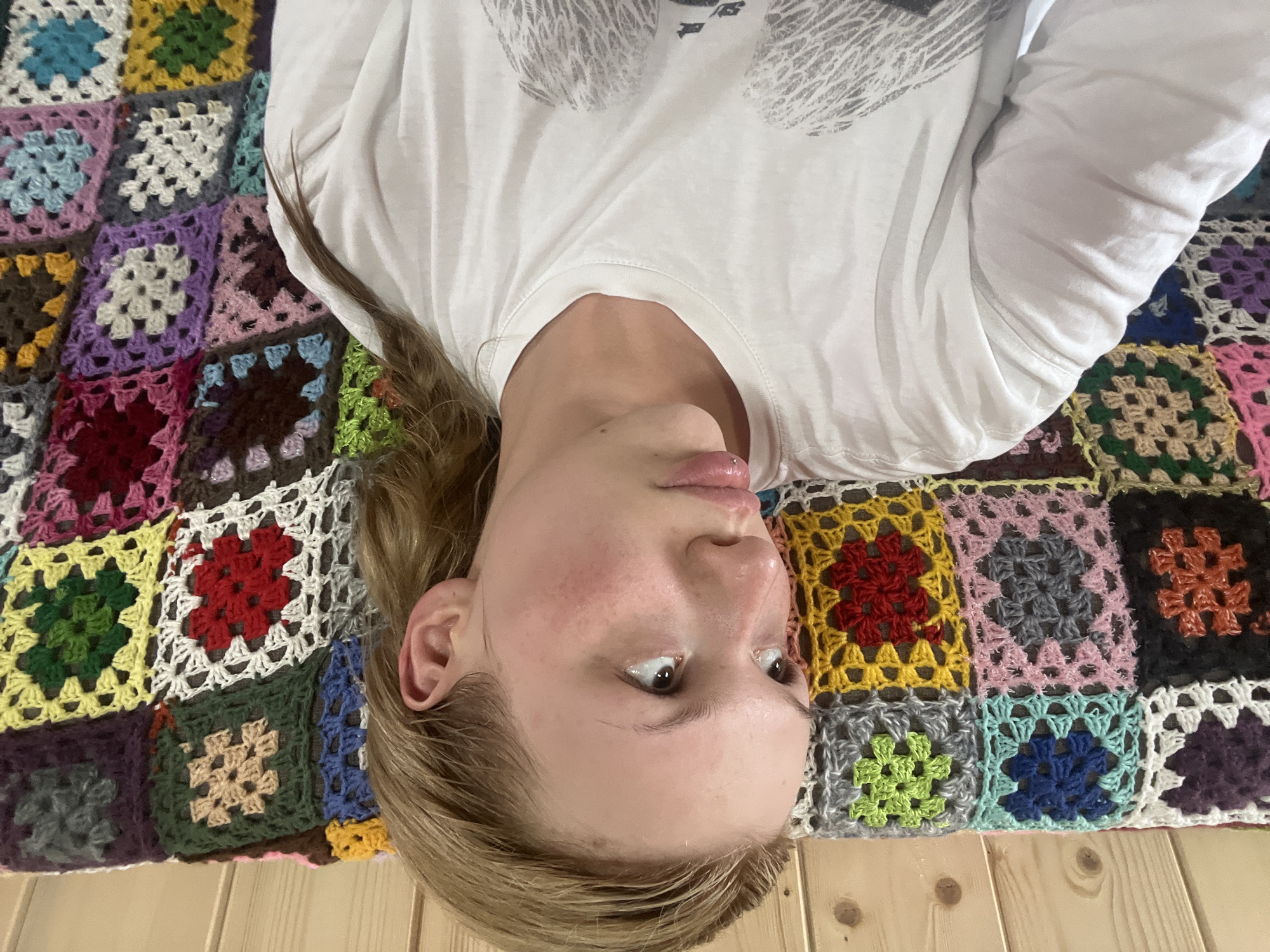 A young person wearing a white long-sleeved shirt is lying on a colorful crocheted blanket, possibly indoors on a light wooden floor. The person is facing upwards in the frame, with their head tilted back. The location cannot be confirmed to be in the city of Chaikovsky, Russia, based on the visual information.