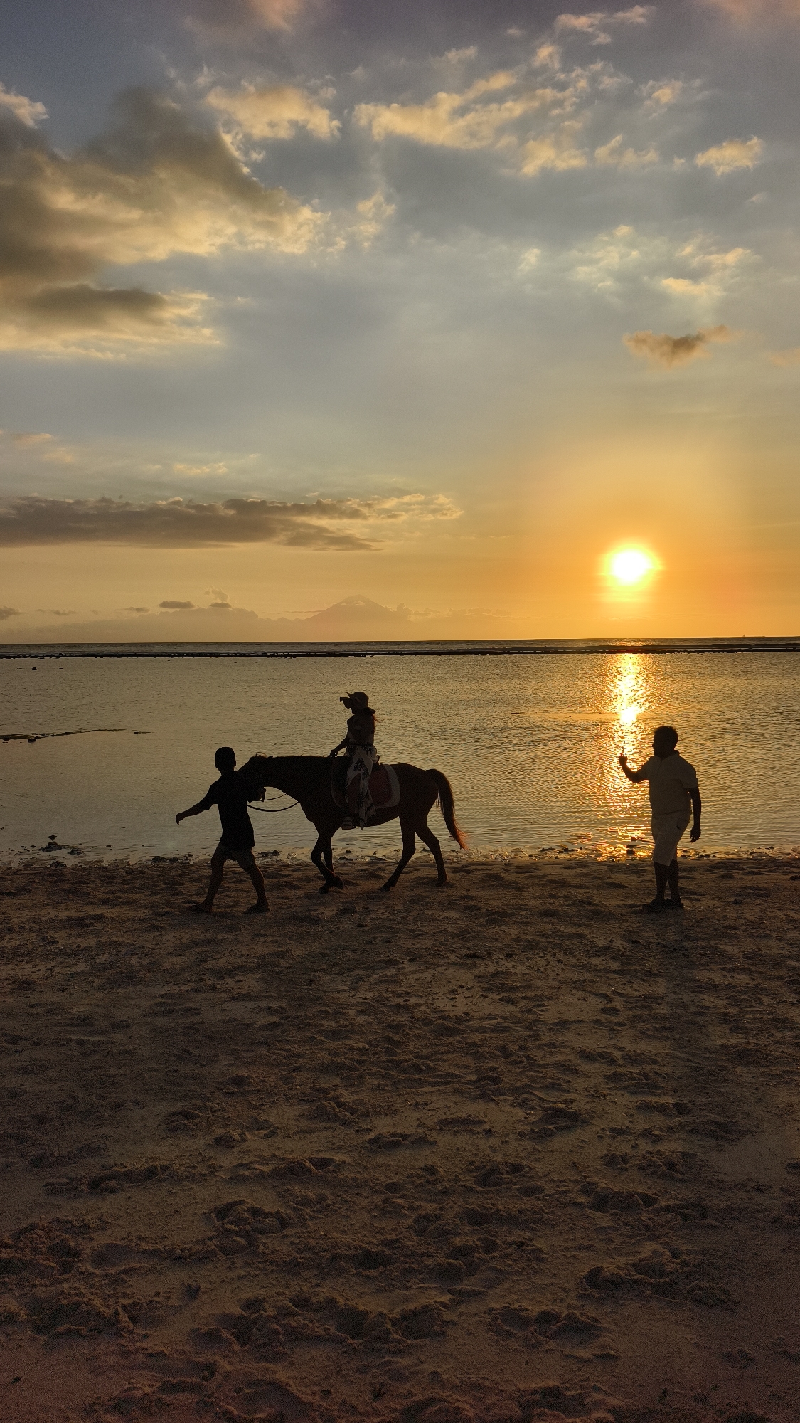 The image depicts a sunset scene on a beach in Unknown, Indonesia. The sun is setting over the ocean, casting an orange glow across the sky and reflecting on the water. Three figures are visible in silhouette. A child sits on a horse led by an adult along the shoreline. Another adult stands in the water, raising their hand in a wave or gesture. In the background, a land mass is slightly visible across the water. The beach is sandy, with darker sand in the foreground. The sky is filled with clouds, some darker and some illuminated by the setting sun.