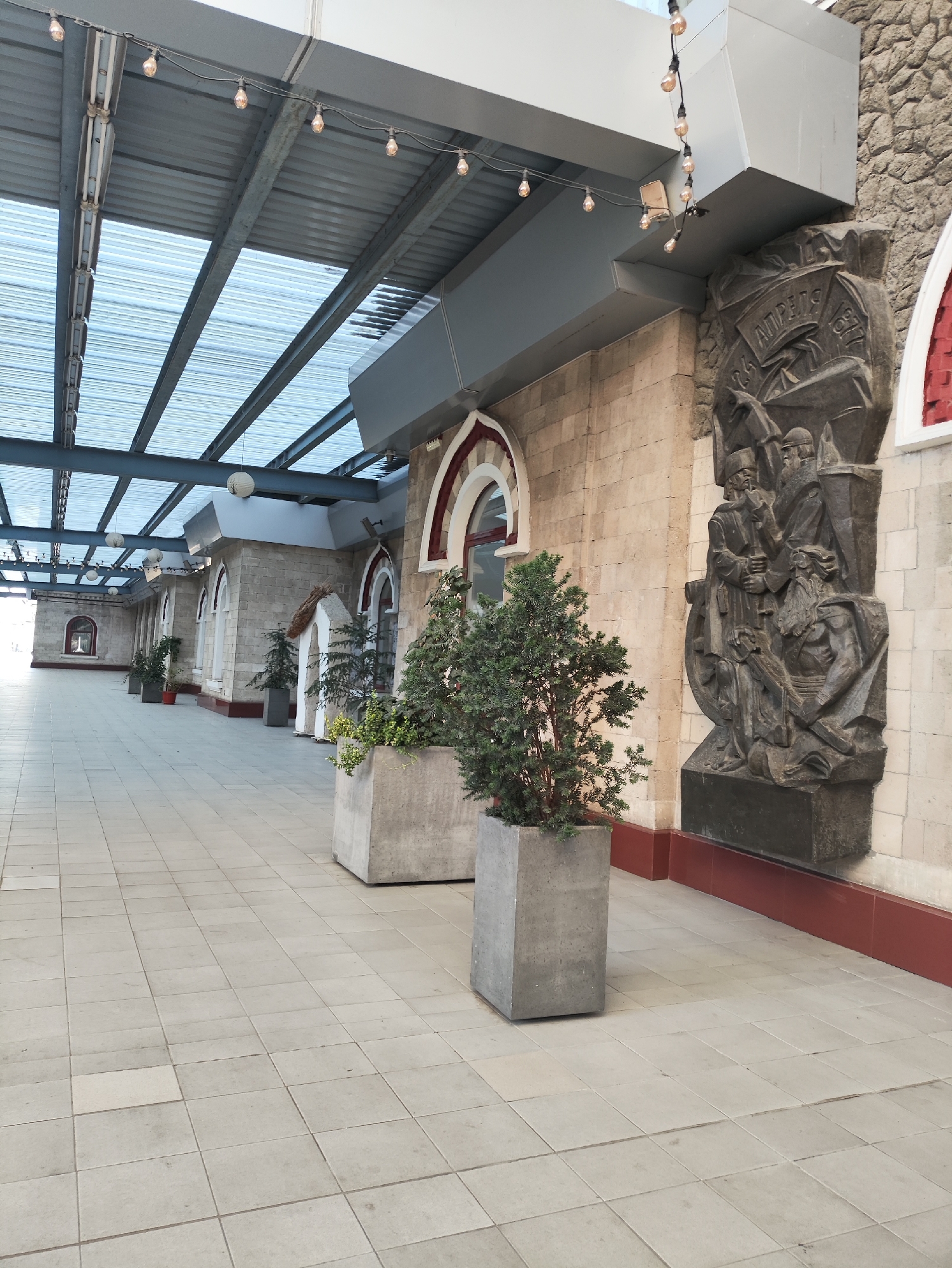 The image shows an outdoor walkway with a covered roof structure made of corrugated metal and glass. Along the walkway, there are several buildings constructed from light-colored stone blocks. These buildings have arched windows with red trim. In the foreground, there are large, square concrete planters containing green bushes. To the right, a large, dark relief sculpture is attached to the stone wall of one of the buildings. The sculpture depicts several figures and text. String lights with Edison-style bulbs are suspended from the roof structure. The walkway is paved with light-colored square tiles. The scene appears to be in a public area, possibly a promenade or an outdoor seating area for a venue. The relief sculpture may be a historical or artistic monument. The overall impression is one of an urban architectural setting with decorative elements.