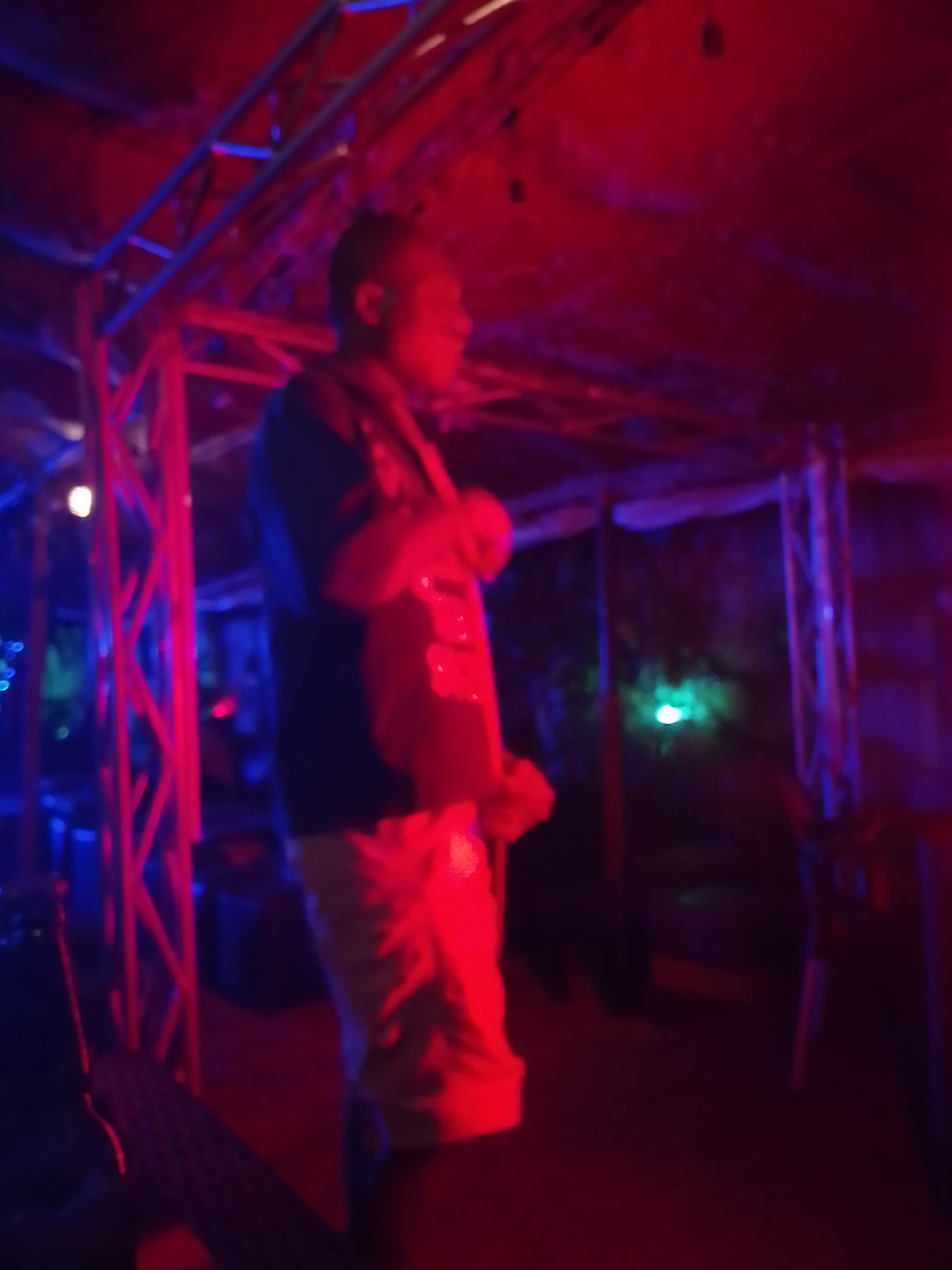 A man stands in an indoor space, illuminated by red and blue lighting, possibly a bar or club. The location is inferred to be Yelwa, Nigeria. The man is wearing a dark-colored shirt and light-colored shorts. A metallic structure is visible behind and to the side of him. Other details are obscured due to blur and the lighting conditions.