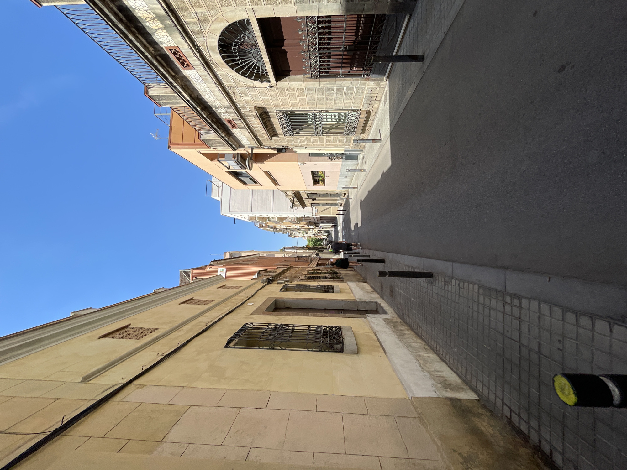 The image depicts a street scene in the Eixample neighborhood of Barcelona, Spain. The street is paved with asphalt and lined with sidewalks made of square tiles. Several black bollards with yellow tops are positioned along the sidewalk on the left side of the street.
On the left side of the street, a building with a pale yellow facade features rectangular windows with ornate iron grills. The building has a stone base and a decorative cornice. Further down the street on this side, more buildings with similar architectural elements are visible.
On the right side of the street, a building constructed of light-colored stone blocks showcases a large arched entrance with a dark wooden door and intricate wrought-iron gates. Above the entrance, decorative patterned stonework and smaller arched openings with iron grills are present. Adjacent to this building, other structures of varying heights and colors, including a peach-colored building with balconies and a white building, line the street.
Two individuals are walking away from the camera on the sidewalk on the left side of the street. One is wearing a dark top and shorts, while the other is wearing a dark top and shorts. The sky is clear and blue, indicating a sunny day.