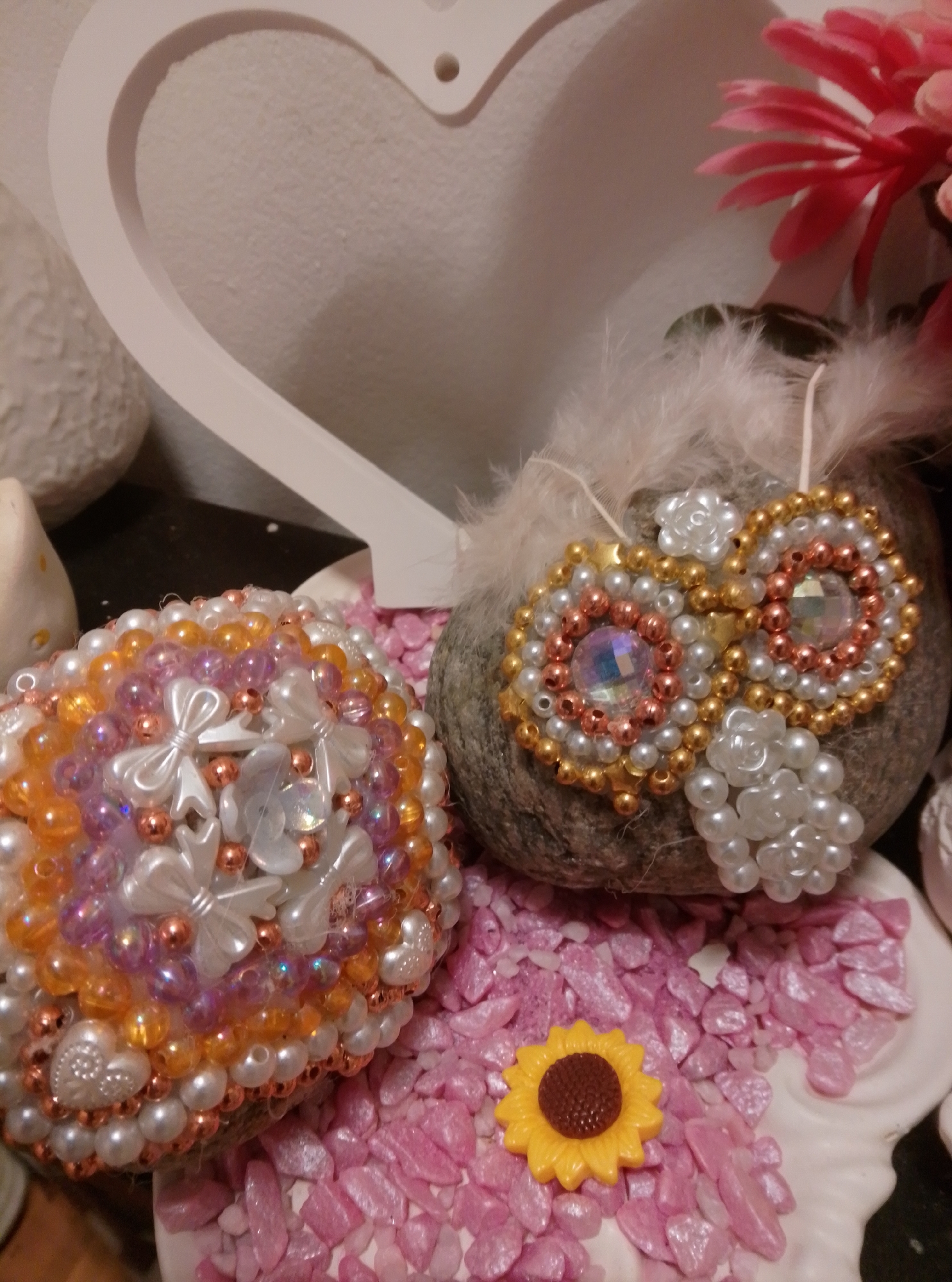 This is a close-up, eye-level, indoor shot of two decorated, spherical objects placed on a surface covered with small, pink, irregular-shaped stones and a white, shell-shaped decorative piece. The object on the left is covered in an intricate pattern of small, round beads in shades of white, pearl, orange, and pink, interspersed with small white bows and heart-shaped embellishments. The object on the right is a grey, pebble-like stone decorated with a symmetrical pattern of gold and white beads, rose gold beads, and two iridescent, multifaceted gems, accented by white beaded flowers and delicate white feathers. In the foreground, a small, yellow plastic sunflower charm rests on the pink stones. In the background, a portion of a large, white, heart-shaped decorative object is visible against a textured, light-colored wall. The lighting is soft and diffused, creating a warm, decorative aesthetic. The image does not provide any information about the city of Wedel, Germany.