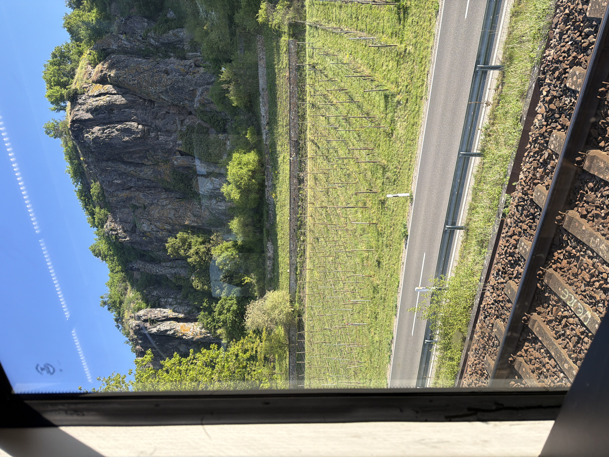 The photograph, taken from inside a moving train near Hüffelsheim, Germany, shows a landscape featuring a steep, rocky hillside partially covered with vegetation. A vineyard with bare vines is visible on a grassy terrace. Adjacent to the vineyard is a road with a guardrail. The train tracks and gravel are prominent in the right side of the frame, as viewed from the window. The sky is clear and blue.