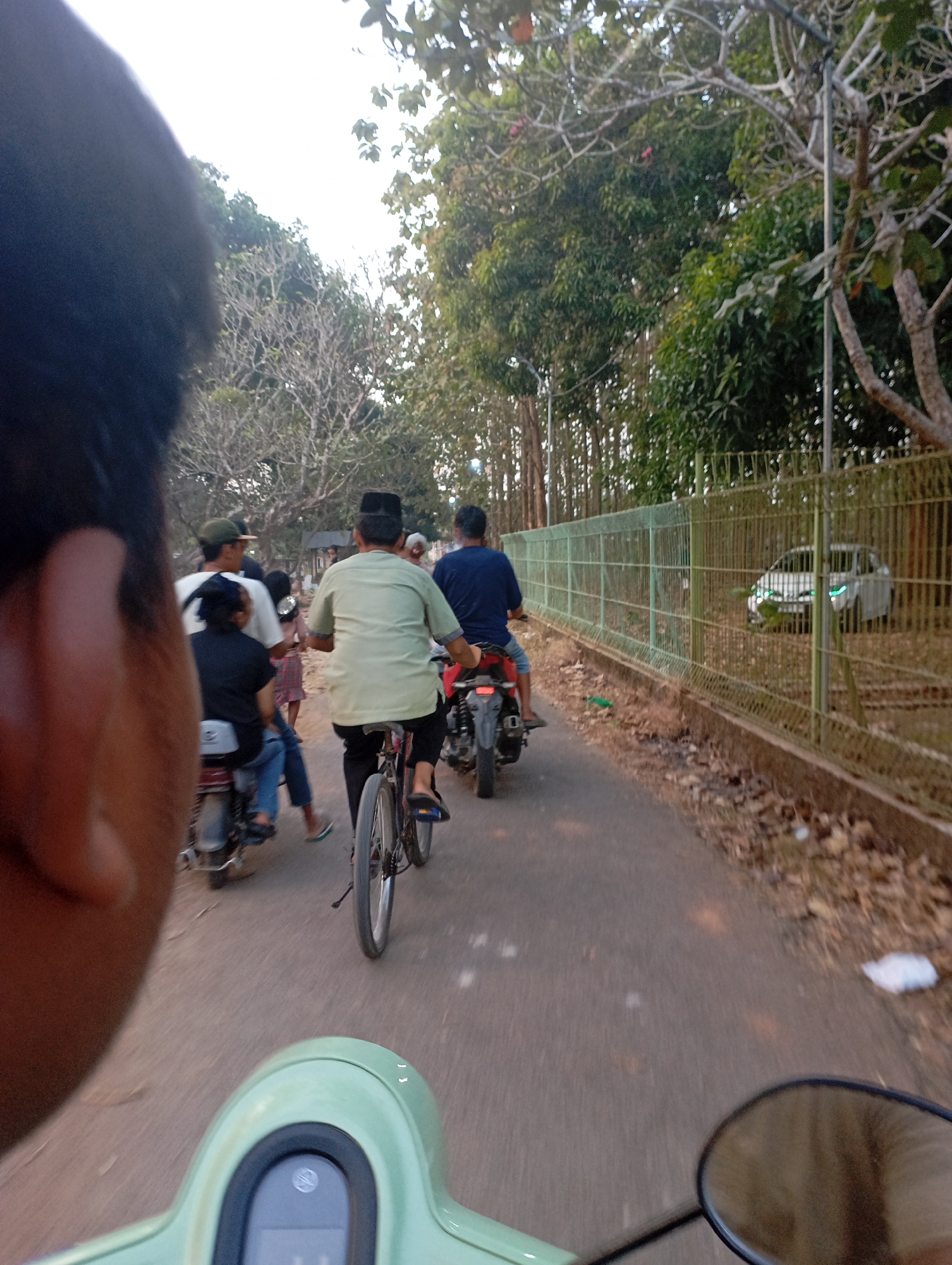 The image depicts a street scene in Kecamatan Haurgeulis, Indonesia, with several people on motorcycles and a bicycle. In the foreground, a person's head and shoulder are partially visible, suggesting they are the photographer or a passenger on a vehicle. Further down the road, a person wearing a light green shirt and a black peci hat is riding a bicycle. To their left, a person is riding a motorcycle with two passengers behind them. In the distance, a white car is parked behind a green metal fence, with trees and vegetation lining the other side of the road. The overall atmosphere suggests a casual daytime activity, possibly a procession or group outing.