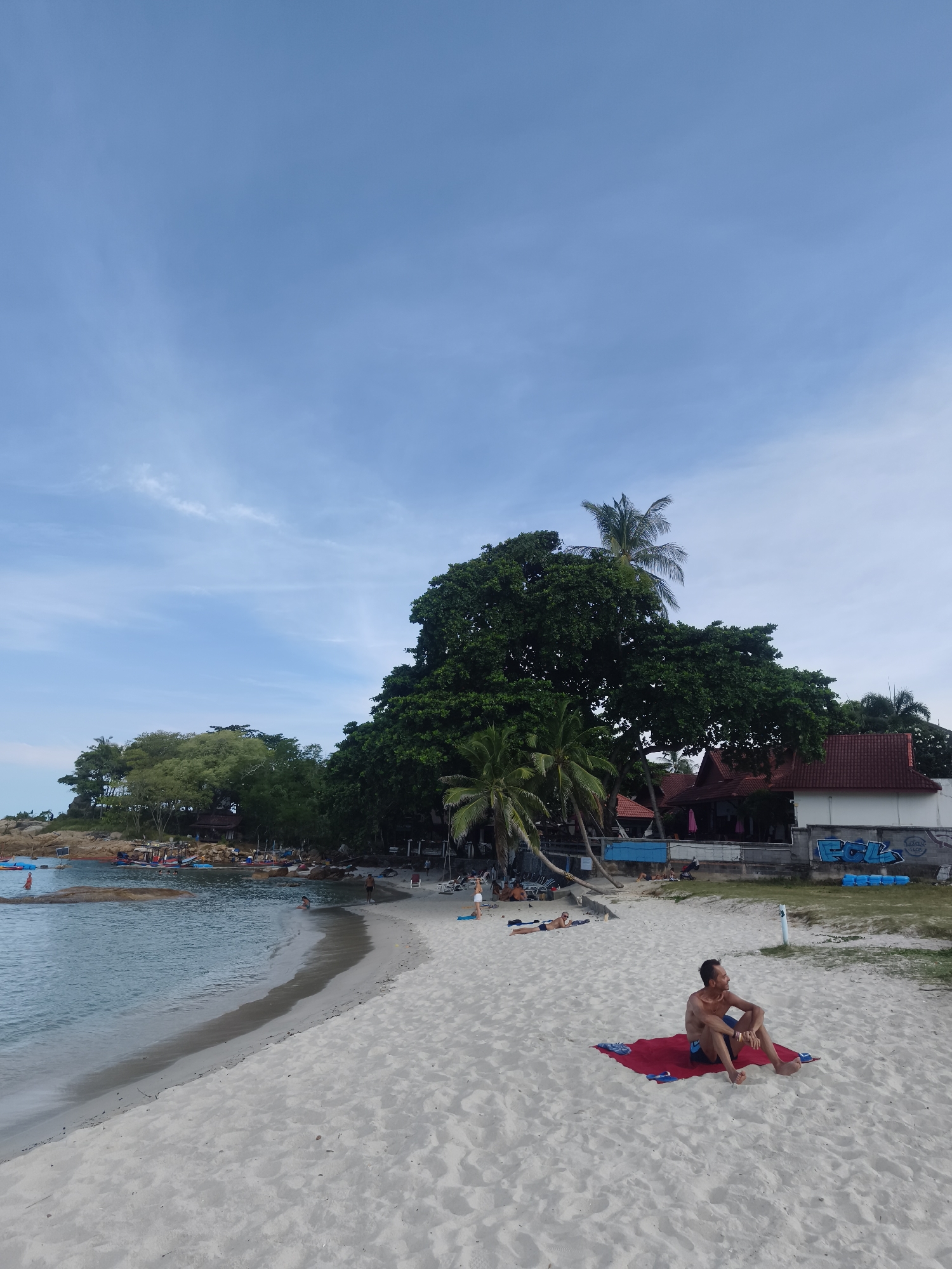 This is a photograph of a beach scene in the Ko Samui District of Thailand. In the foreground, a man is sitting on a red towel on the white sand beach, looking to his right. He is wearing blue swim trunks and is shirtless. Behind him, other beachgoers are visible, some sunbathing on chairs and towels. The beach curves to the left, meeting the calm, turquoise water. In the background, lush green trees, including palm trees and a large, full-canopied tree, dominate the landscape. Several buildings with red roofs are situated among the trees, and some boats are moored in the water near a rocky outcrop. The sky above is a pale blue with scattered white clouds. The scene depicts a relaxed tropical beach environment.