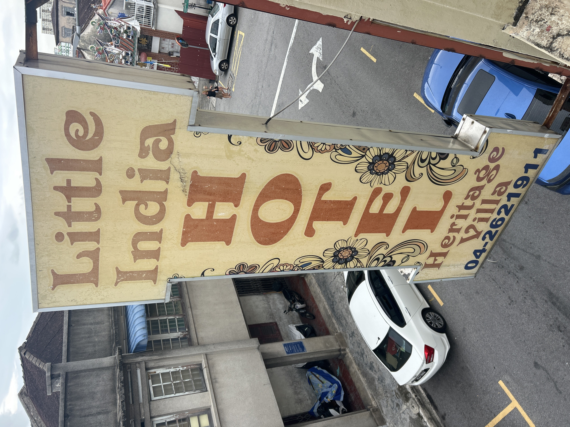 The image shows a close-up view of a weathered sign for "Little India Hotel Heritage Village" in George Town, Malaysia. The sign is yellow with brown lettering and features floral decorative elements. Below the sign, a street is visible with parked cars, including a white sedan and a blue car. A motorcycle is parked under an awning in an alleyway to the left. The architecture suggests an older urban environment. The sky is overcast.