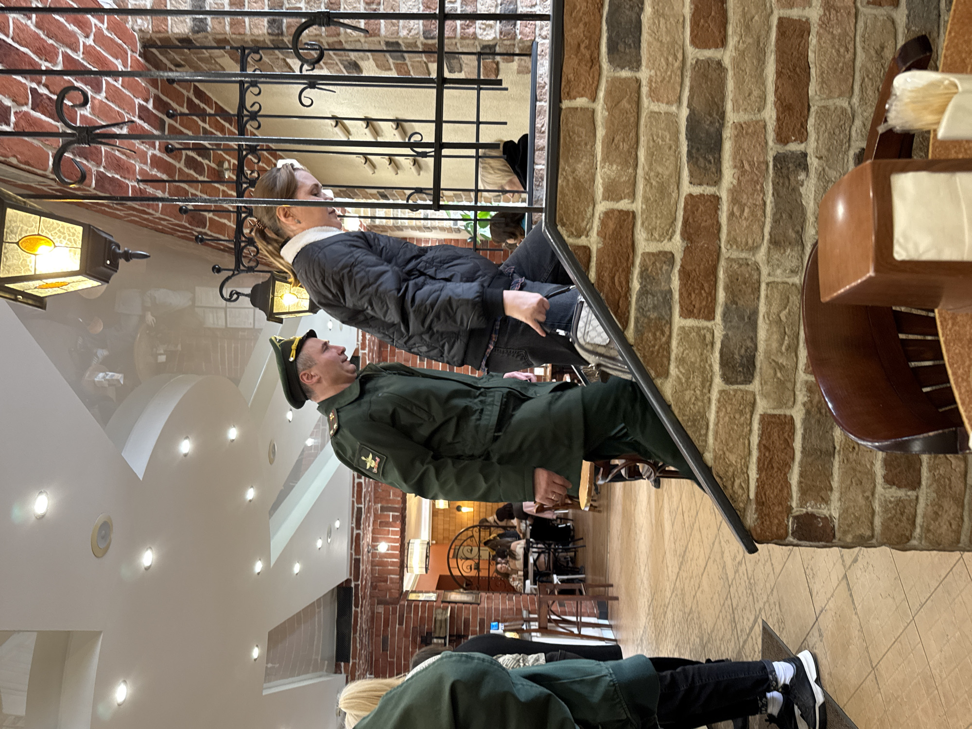 Inside a building in Saint Petersburg, Russia, a man in a green military uniform and hat stands near a woman with a black jacket and jeans near a black metal railing and brick wall. The interior reveals tables and chairs with people seated, a decorated metal partition, and overhead lighting. Another woman is seen nearby.