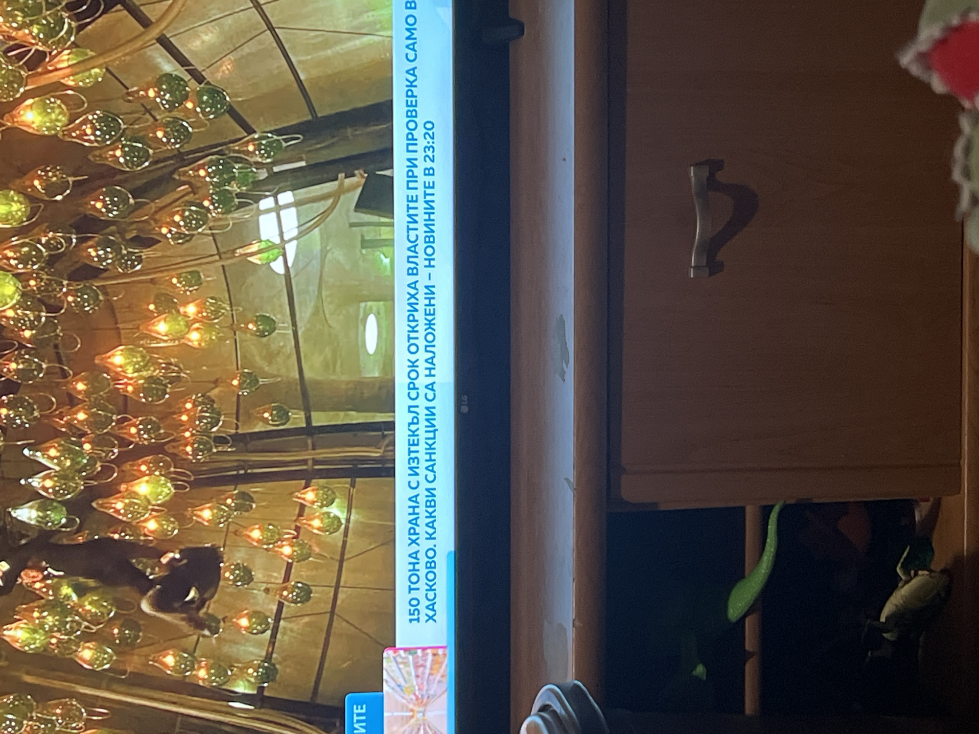 The image depicts a television screen displaying a program, positioned atop a wooden media console in an indoor setting.
**Television Content:**
The television, an LG brand unit (indicated by the logo on the bottom frame), displays an interior scene with a high ceiling heavily adorned with numerous illuminated, teardrop-shaped glass luminaires. These lights glow predominantly in green and yellow-orange hues. A dark-clad human-like figure is observed hanging upside down from the ceiling structure in the upper-left portion of the screen.
Below the visual content, a horizontal blue news ticker is displayed. The Bulgarian text translates to: "150 TONS OF EXPIRED FOOD WERE DISCOVERED BY AUTHORITIES DURING AN INSPECTION ONLY IN HASKOVO. WHAT SANCTIONS HAVE BEEN IMPOSED - NEWS AT 23:20."
**Physical Environment:**
Below the television is a light brown wooden media console or dresser. The left side of the console features an open, dark compartment where a bright green, serpentine object (resembling a toy snake) is visible draped over an interior shelf, along with other indistinct dark items at the bottom. The right side of the console consists of a closed drawer equipped with a single, dark-colored, curved handle. A light-colored, partially visible cylindrical object is present on the top surface of the console, to the left.
**Location:**
Based on the Bulgarian language news ticker, the image was captured in Bulgaria. While the specific request implies a location within Sofia, Bulgaria, the visual information provided in the image (a generic interior, home furniture, and a movie/TV show scene on the screen) does not contain any identifiable landmarks or specific geographical markers to pinpoint a precise location within the city of Sofia. The content displayed on the television is a fictional or movie setting, not a real location in Sofia.