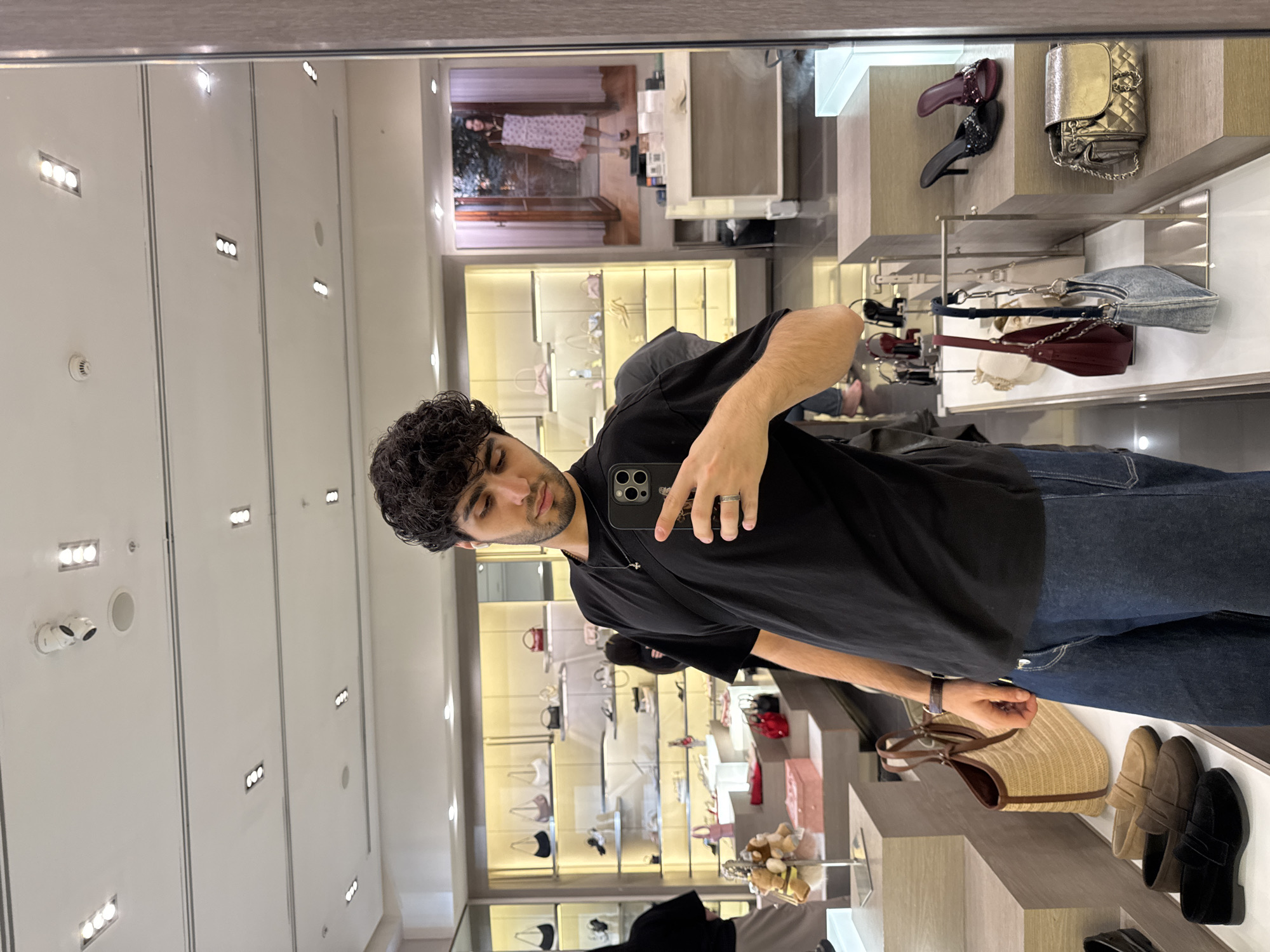 A man is taking a selfie in front of a mirror inside a store. The store displays handbags and shoes, showcasing various brands and styles. The location is possibly in Yerevan, Armenia, judging by the general ambiance. The man is wearing a black shirt and blue jeans. The background features shelves filled with merchandise. The lighting is bright and even, suggesting a well-lit retail environment.
