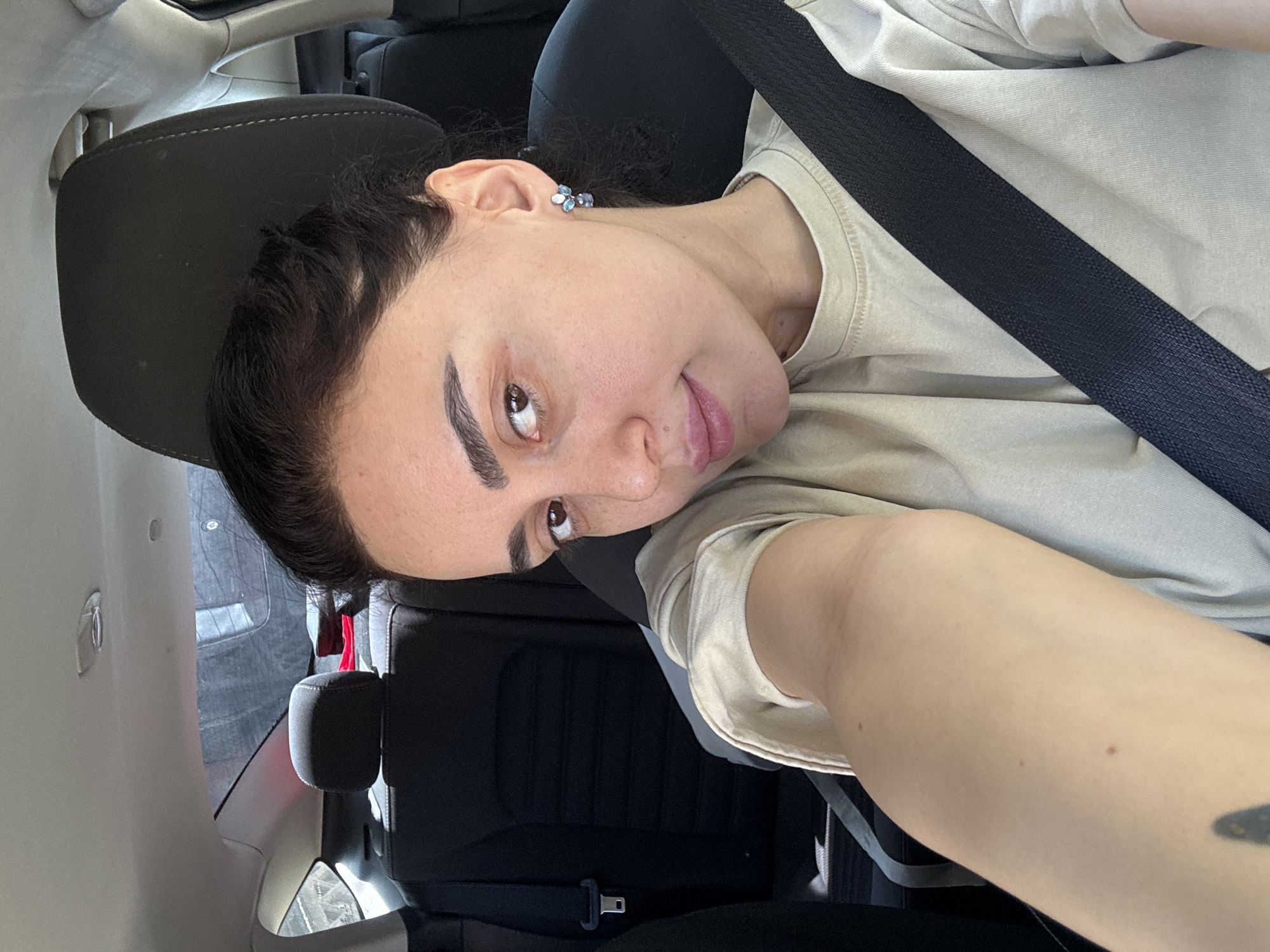 A person with fair skin and dark hair is in a car, wearing a seatbelt and a beige t-shirt. The person has distinct eyebrows and is wearing a light blue earring. The interior of the car is visible, including the black seat and headrest. The location is likely Tbilisi, Georgia, but this cannot be confirmed from the image alone.