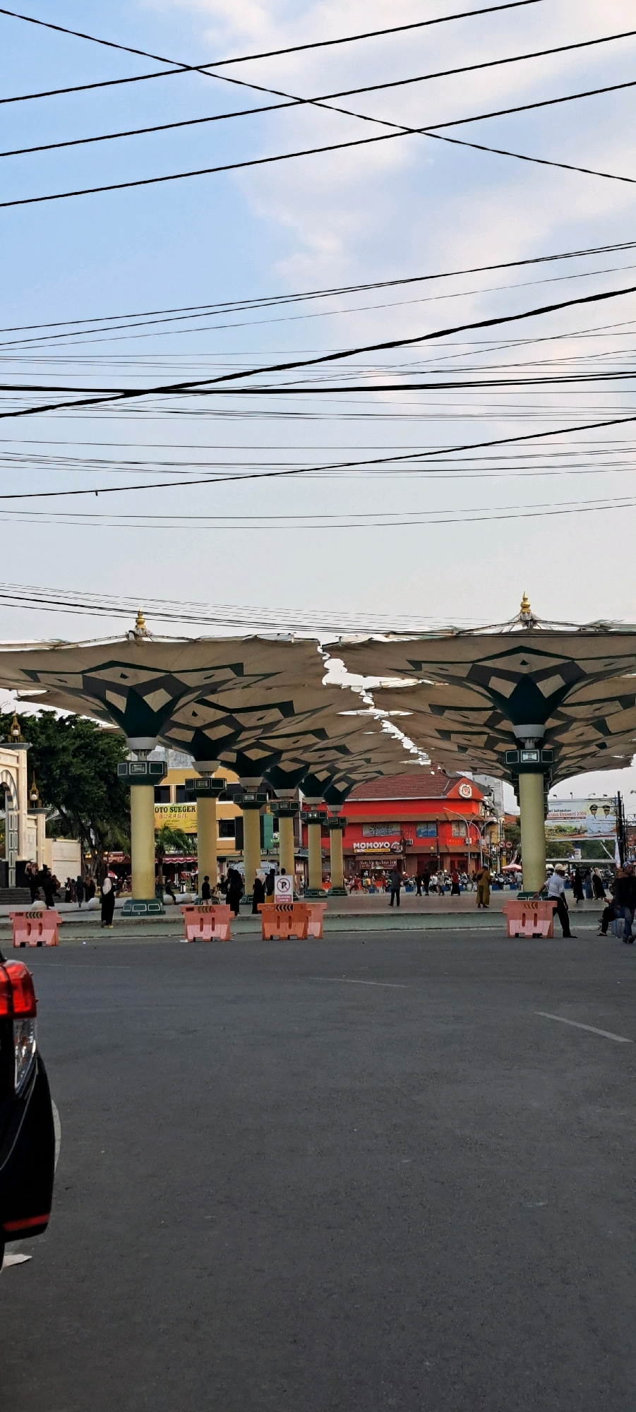 The image shows a street scene in Pasuruan, Indonesia, characterized by a large, white, patterned canopy supported by columns. The canopy appears to be made of fabric and is partially torn in places. It spans across a paved road where several cars and people are visible. The background features buildings, including a red building with signage that reads "MOMOYO". Other structures include a white building with arched windows, a yellow building with signs, and a billboard. The sky is visible above, with a network of overhead electrical wires crisscrossing the frame. Several orange and white traffic barriers are placed on the road. People are walking and standing in various areas, some appearing to be tourists or locals. The lighting suggests it is daytime, likely late afternoon, with a bright sky.