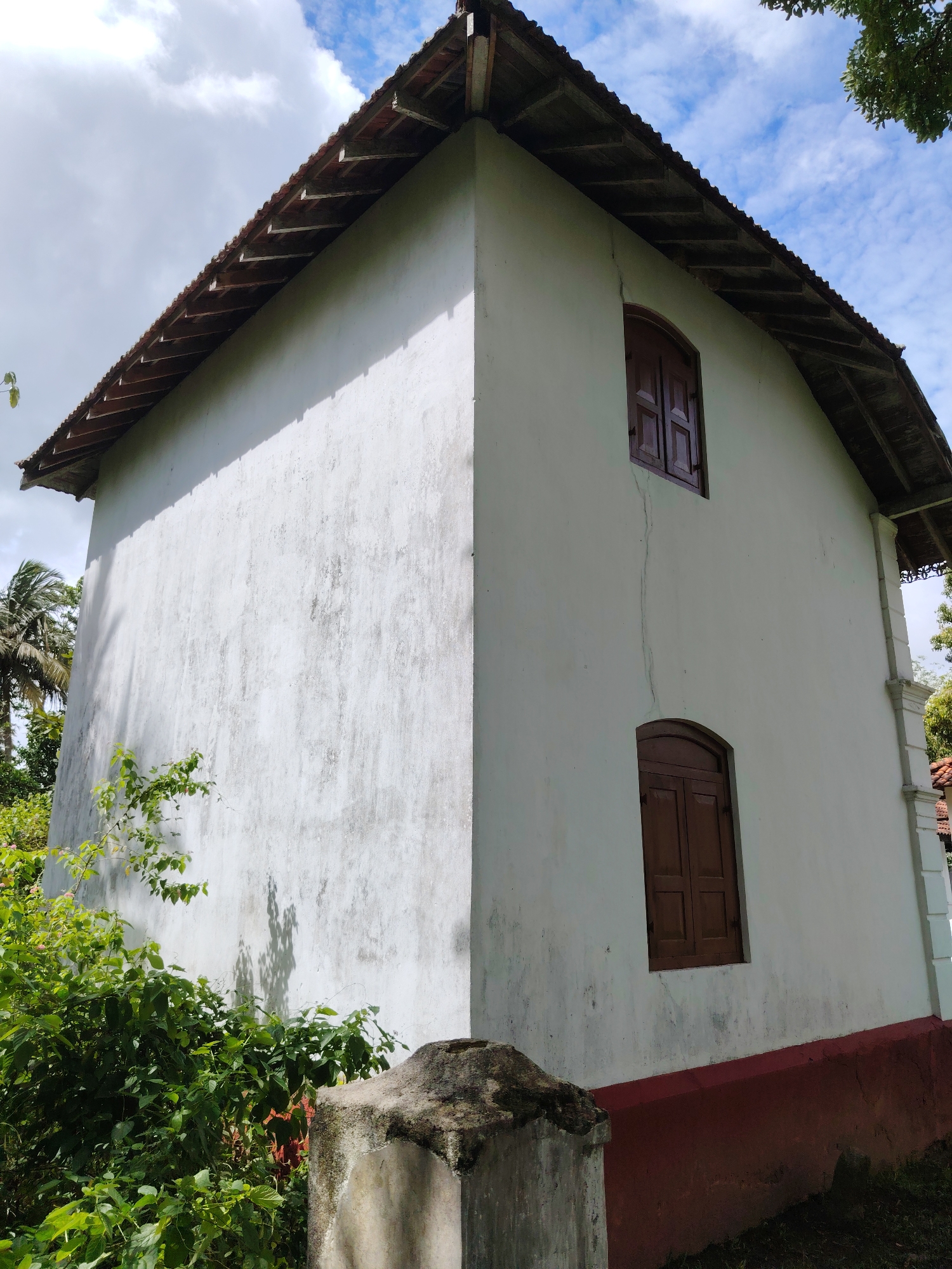A low-angle shot captures the corner of a white, two-story building under a blue sky dotted with white clouds. The building has a weathered white stucco exterior with a prominent vertical crack running down the wall between two windows on the right side. The lower part of the building's exterior wall is painted a contrasting reddish-brown.
The roof is made of reddish-brown tiles, with exposed wooden rafter ends visible beneath the eaves, casting small shadows. Two closed wooden windows are visible on the right face of the building; the lower one has an arched top, while the upper one is rectangular. Both windows feature dark brown louvered shutters.
Lush green foliage and bushes grow around the base of the building, partially obscuring the lower section of the wall on the left side and in the immediate foreground. Several palm trees are visible in the background on the left, suggesting a tropical or subtropical setting. A weathered concrete or stone structure is partially visible in the bottom right foreground. The lighting indicates it is daytime, likely late morning or early afternoon, with bright sunshine. There is no visible text or human activity.