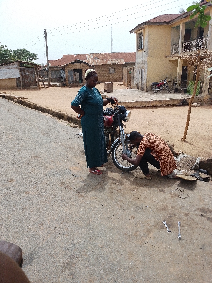 The image depicts an outdoor scene in Igbo Ijaiye, Nigeria, where a motorcycle is undergoing repair on an unpaved shoulder adjacent to a paved road.
**Subjects:**
* A female adult stands to the left of the motorcycle, facing right, looking towards its front wheel. She wears a dark green, patterned long dress, a patterned head covering, and red flip-flops. Her left hand rests on the motorcycle's handlebar, and her right hand is on her hip.
* A male adult is crouched on the dirt ground to the right of the motorcycle, engaged in repairing or inspecting the front wheel assembly. He wears a patterned orange and black long-sleeved shirt and dark trousers. His hands are actively manipulating the motorcycle's front wheel area.
**Objects:**
* A red and silver motorcycle is the central subject, positioned on the dirt ground. Its front wheel is being serviced.
* Two silver open-end wrenches are lying on the asphalt road surface in front of the crouching male. Additional tools and a piece of fabric or mat are visible on the dirt near his position.
* In the background, a second red motorcycle is parked near a building.
**Environment:**
* The immediate foreground shows a section of gray asphalt road, transitioning to a lighter-colored dirt or sandy ground beyond its edge.
* The background features several buildings:
* To the left, multiple single-story structures with corrugated metal roofs and walls made of what appear to be unpainted bricks or blocks.
* To the right, a two-story building with a light beige/yellow facade and two balconies with metal railings.
* A wooden utility pole with multiple overhead wires is visible in the mid-ground against a bright, hazy sky.
* Sparse vegetation is present in the distant background.