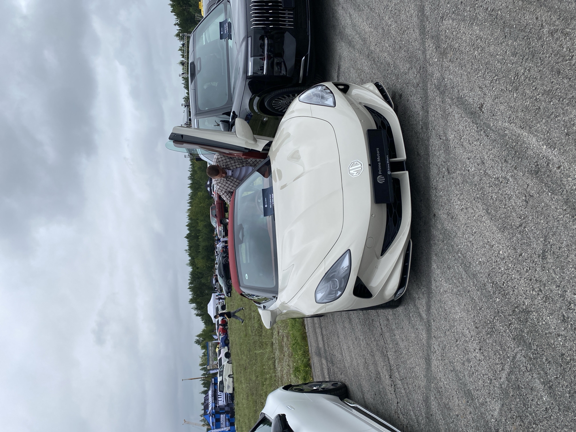 This image depicts an outdoor event, possibly a car show, with several vehicles present. In the foreground, a cream-colored convertible sports car with a red top is prominently featured. Its doors are open, with one of them a gull-wing style door. A person in a plaid shirt is leaning into the sports car. To the right of the sports car is a large black SUV. In the background, there are various tents, other cars, and a line of trees against a cloudy sky. There are also people walking around and some event infrastructure, such as a stage or booth setups, visible. The ground is paved with asphalt. The presence of multiple vehicles and tents suggests a public gathering, likely related to automotive interests. The image does not contain any identifiable landmarks or clues that would pinpoint the exact location as Perm, Russia.