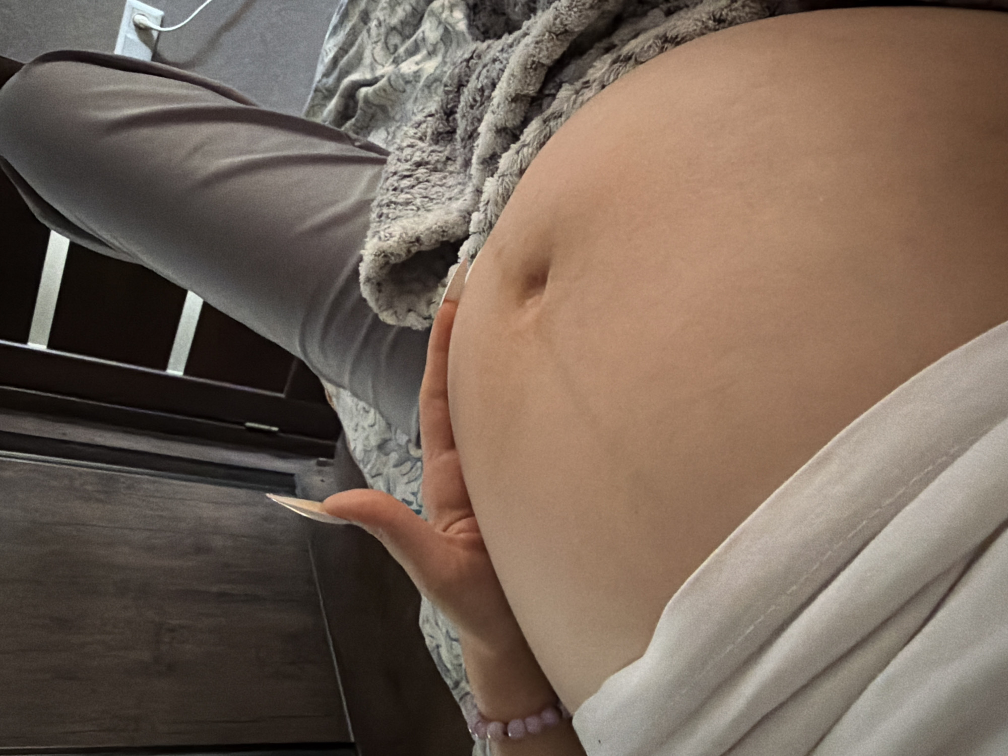 The image shows a close-up view of a pregnant person's abdomen, with a hand resting on it. The person is lying down, as indicated by the presence of a blanket and a leg covered by grey pants. The background appears to be an indoor setting, possibly a bedroom, with a wooden wardrobe visible. There is an electrical outlet with a cord plugged in, suggesting a modern living space. The overall lighting is soft and diffused, creating a warm and intimate atmosphere. There is no information available to suggest the location is in Tbilisi, Georgia.