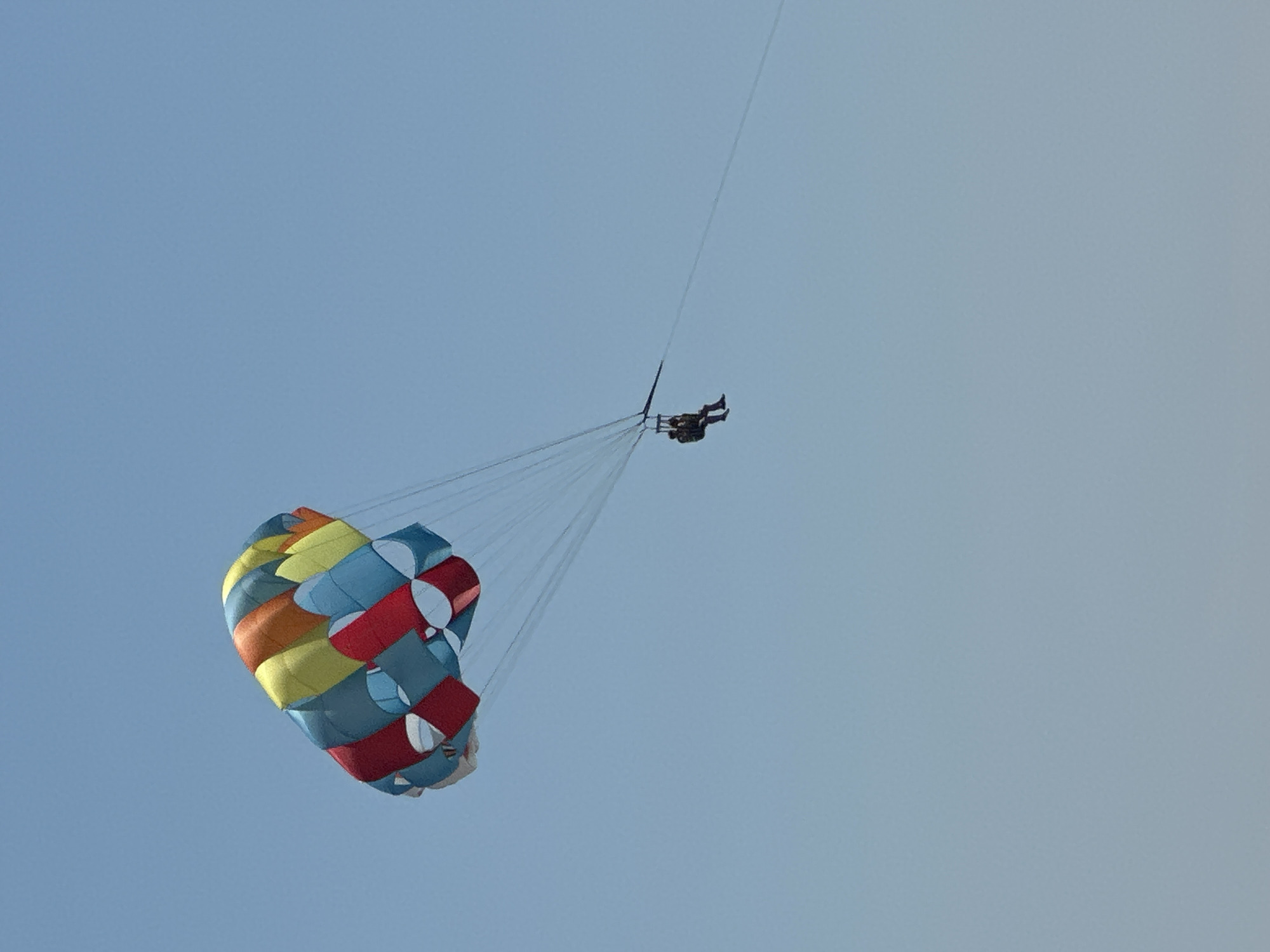 In the sky above Boreti, Montenegro, two individuals are parasailing. A large, brightly colored parachute, featuring panels of red, yellow, orange, and blue, is suspended in the clear blue sky. Attached to the parachute by a series of ropes is a harness, from which two people are seated, facing forward. The parasail and its occupants are ascending into the sky, with the parachute taut and full of air. The background is a uniform, cloudless blue sky.