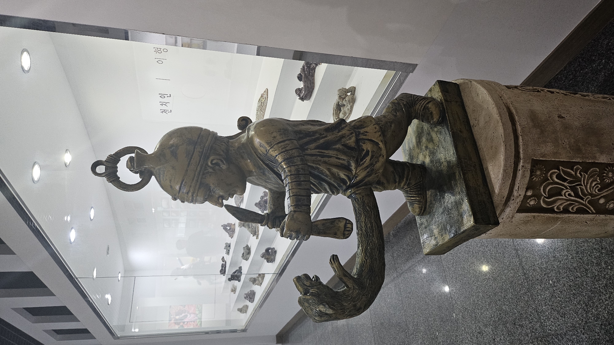This image shows a bronze statue displayed in a museum in Seogwipo-si, South Korea. The statue depicts a male figure wearing a helmet and a tunic, holding a dagger in his right hand. He is standing with his left arm extended, and a carved animal's head is attached to it, biting into a large phallus that emerges from the figure's lower body. The statue is placed on a pedestal in what appears to be a gallery with display cases in the background. The display cases contain smaller artifacts. There is Korean text visible on a white wall in the background, which appears to be part of an exhibit label. The floor is a speckled gray material, and the lighting suggests an indoor exhibition space.