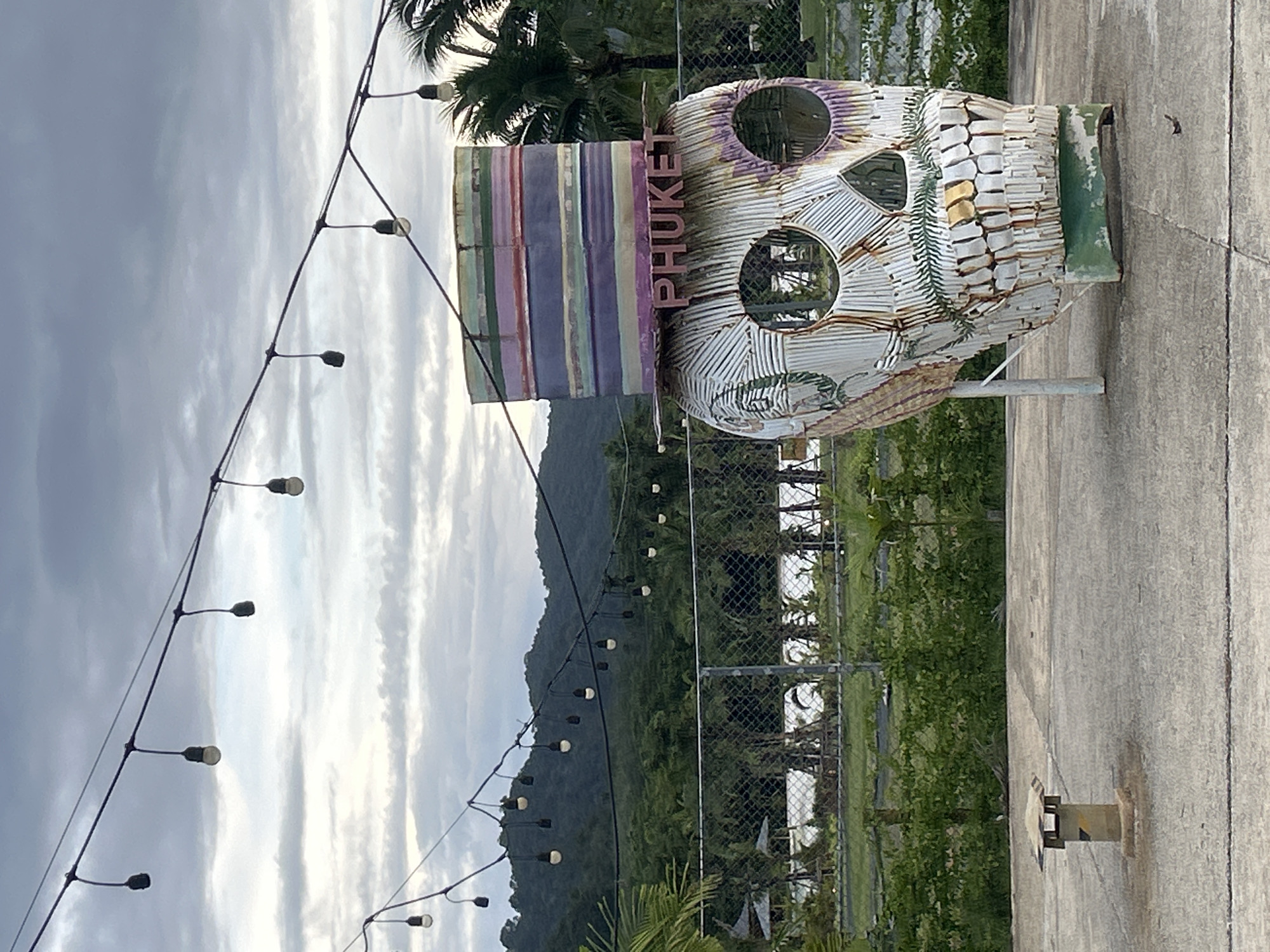 An outdoor scene in Choeng Thale, Thailand, captures a large, stylized skull sculpture under an overcast sky. Numerous strands of string lights with visible bulbs crisscross the upper portion of the frame, some appearing illuminated. In the background, dark green, densely forested hills rise under the grey-white cloud cover. A chain-link fence spans the mid-ground, behind which a body of water reflects the sky, bordered by vegetation and distant small structures.
The main subject, positioned on the right, is a towering skull sculpture made from corrugated metal. It is primarily painted white with rusted accents, featuring large, round, hollow eye sockets and a detailed jaw with teeth. A multicolored top hat, striped horizontally in shades of purple, pink, green, and red, rests atop the skull. The word "PHUKET" is prominently displayed in raised, reddish-brown letters across the skull's forehead. A pale support pole is visible behind the skull's left side.
The foreground consists of a grey concrete or paved surface with visible seams. A short, square-topped fixture, possibly a vent, stands on the left side of the concrete. Sparse green foliage, including palm fronds and bushes, borders the concrete and fills the mid-ground and distant areas. No individuals are present in the scene.