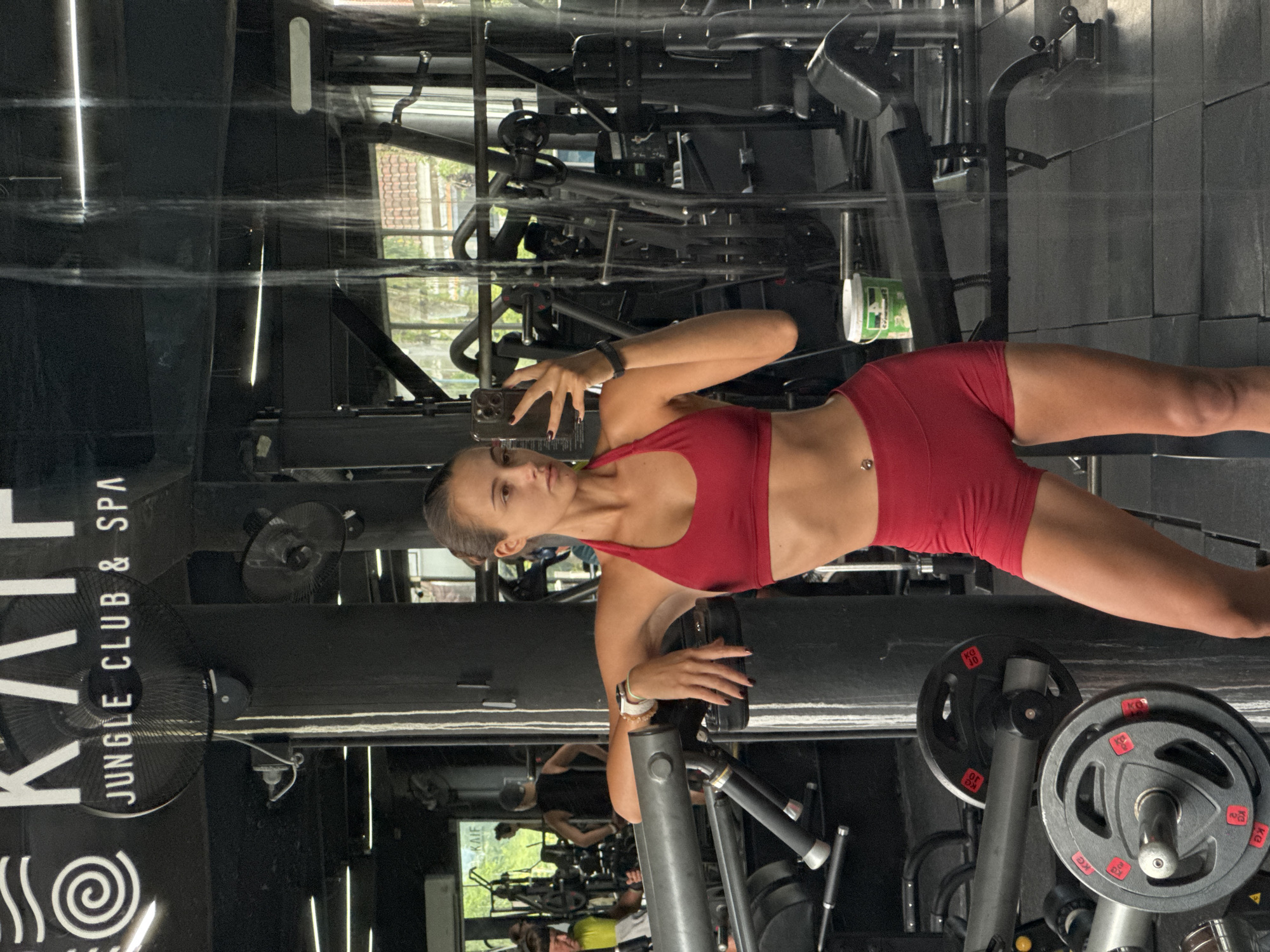 A woman in a red workout outfit is standing in a gym in Kathu, Thailand, taking a selfie in front of a weight machine. The gym has dark walls and black equipment. The name "KAIF JUNGLE CLUB & SPA" is visible on a sign. Other people can be seen in the gym's background.